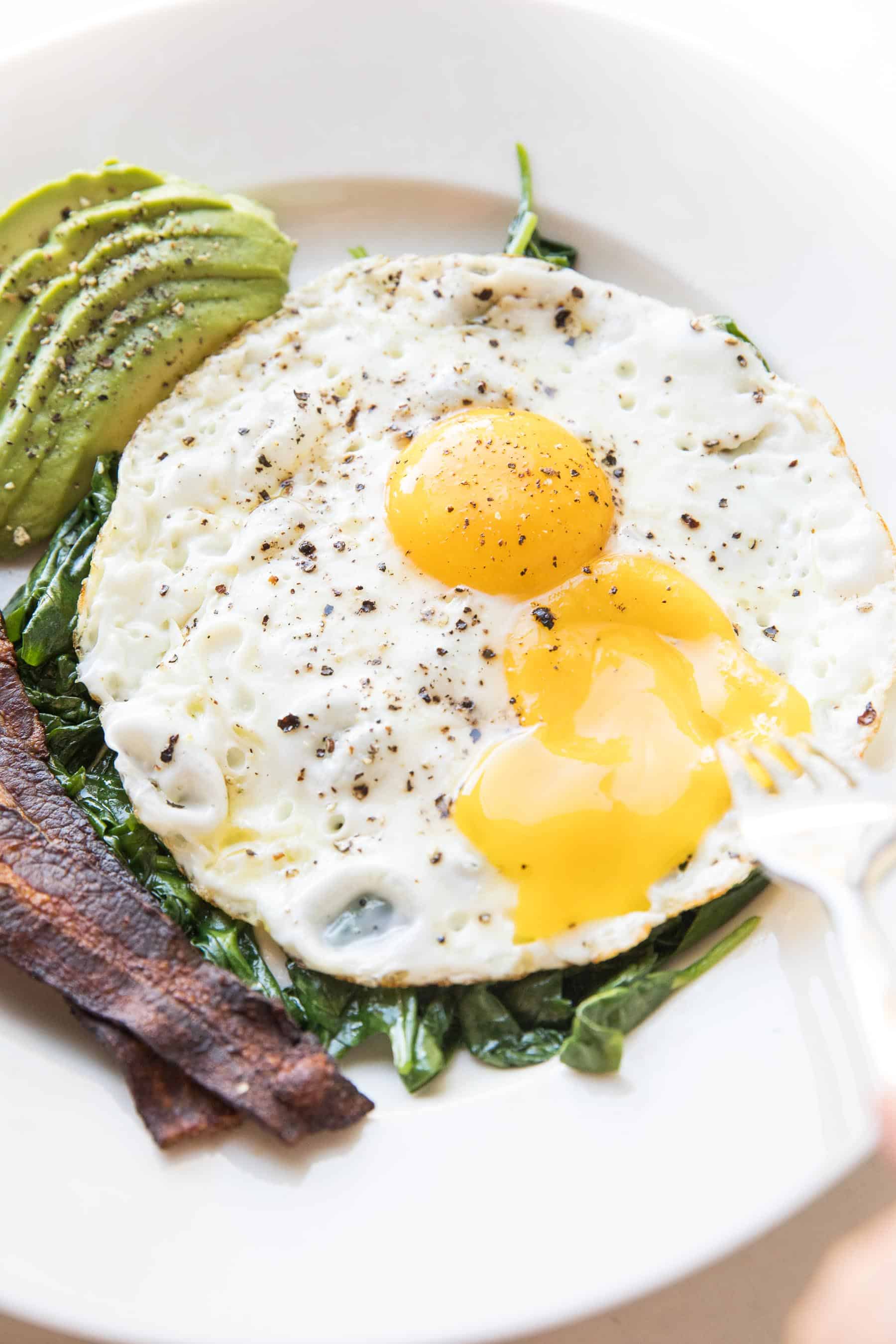 fork breaking sunny side up eggs with sauteed spinach, bacon + avocado