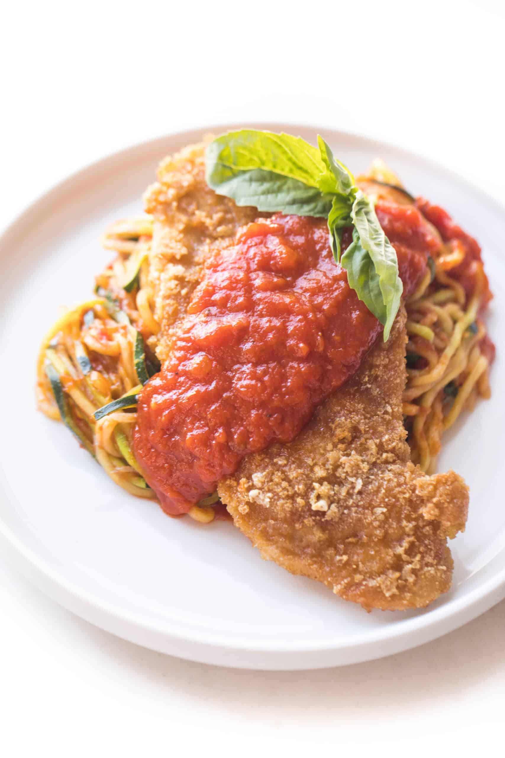 chicken parmesan topped with marinara sauce over zucchini noodles on a white plate and background