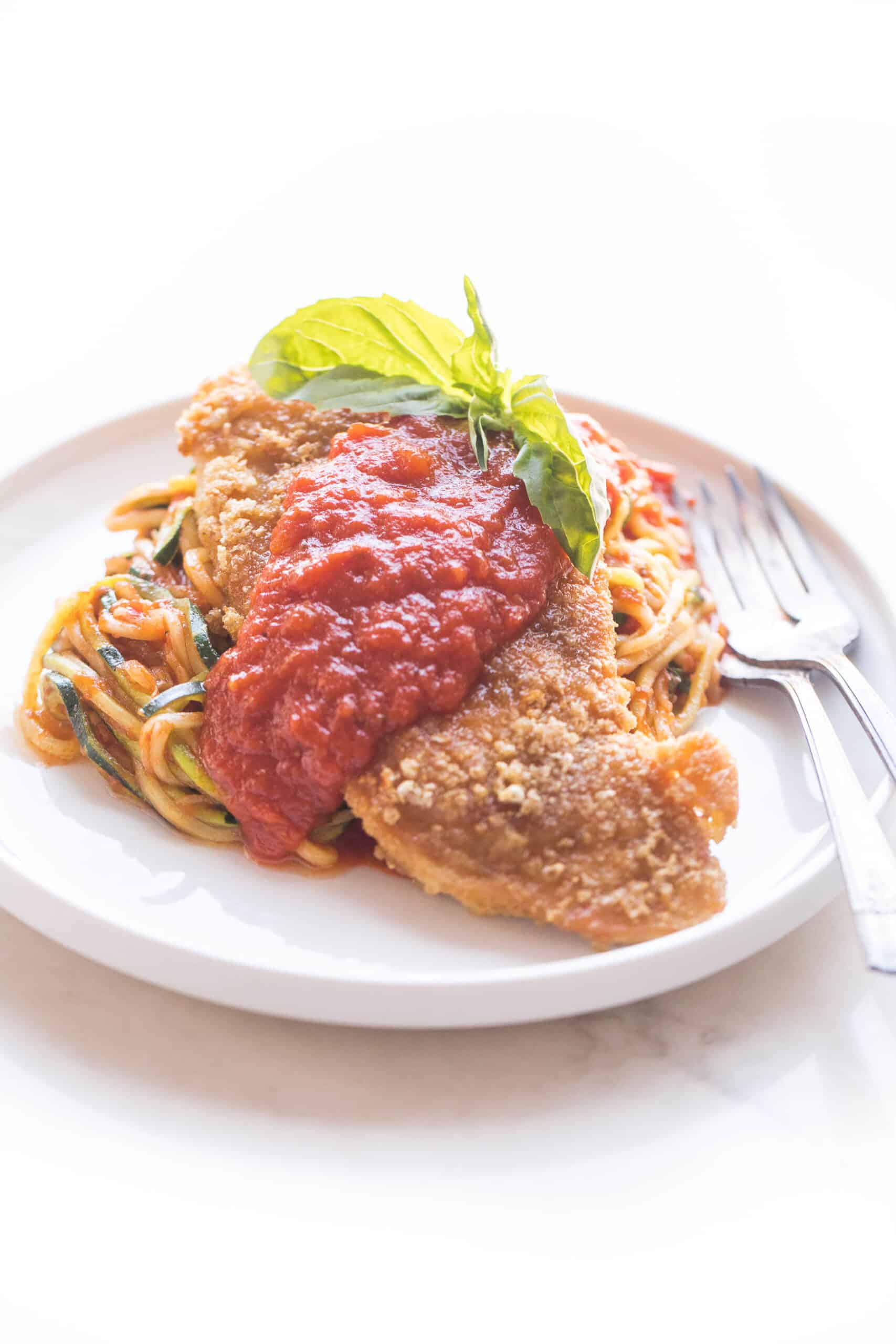 chicken parmesan topped with marinara sauce over zucchini noodles on a white plate and background