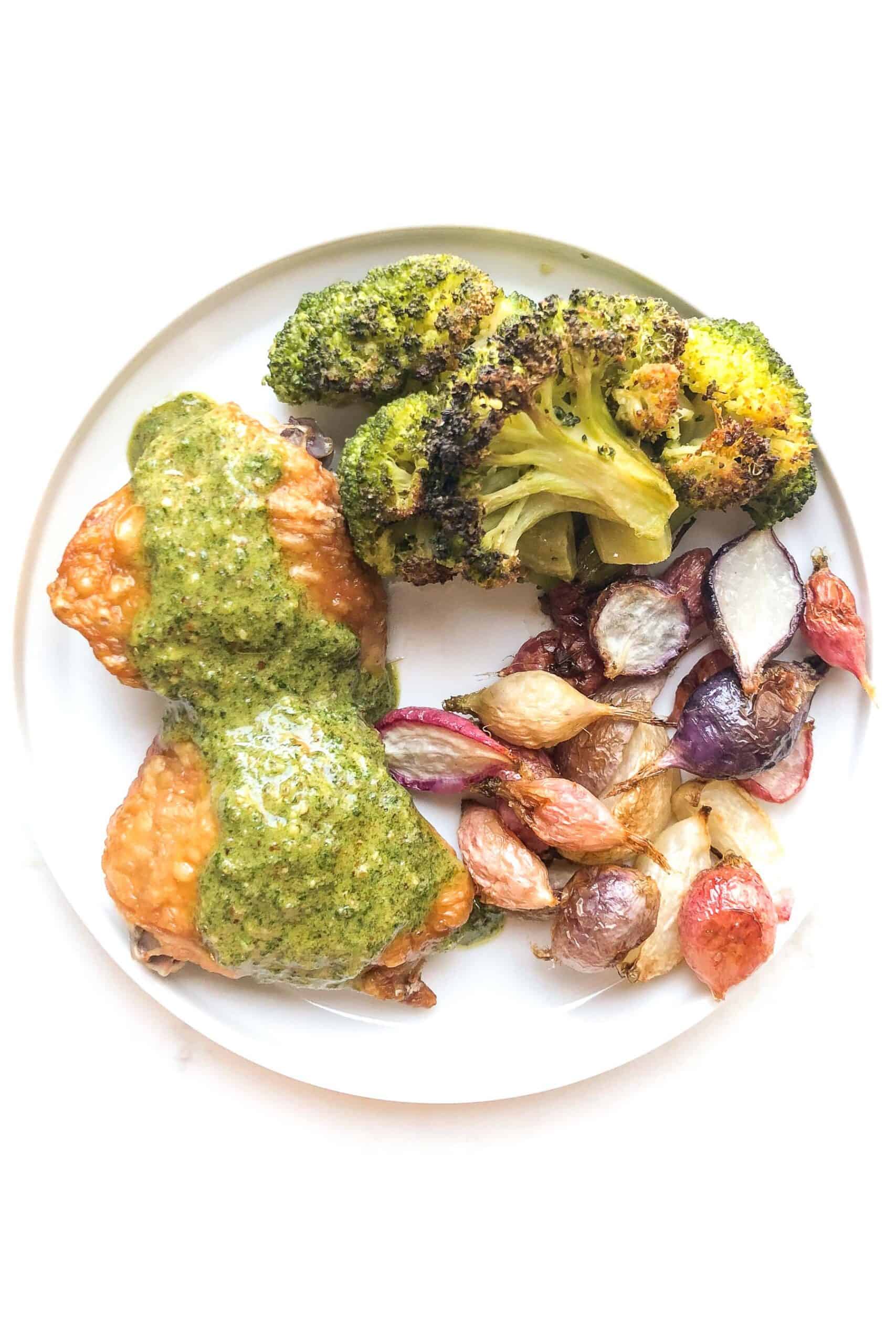 crispy chicken thighs topped with a green orange parsley sauce, roasted radishes and roasted broccoli on a white plate and background