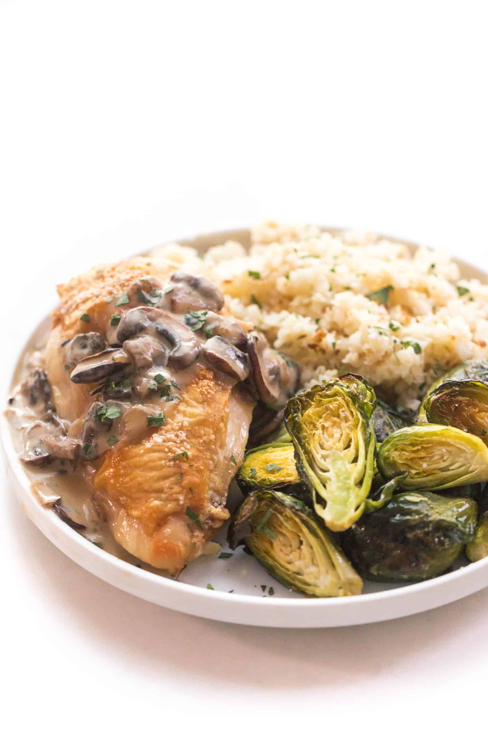 chicken with mushroom gravy on a white plate and background with roasted brussels sprouts and cauliflower rice