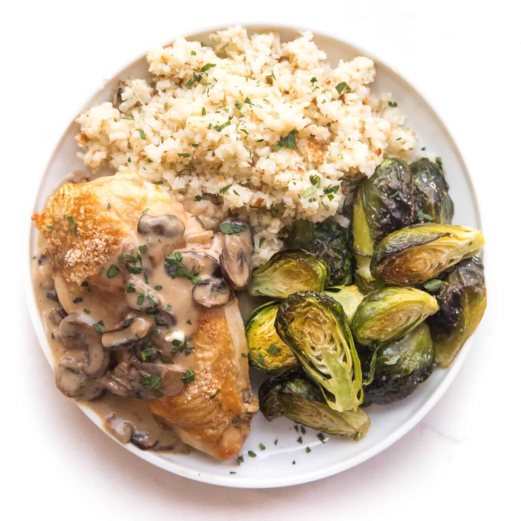 chicken with mushroom gravy on a white plate and background with roasted brussels sprouts and cauliflower rice