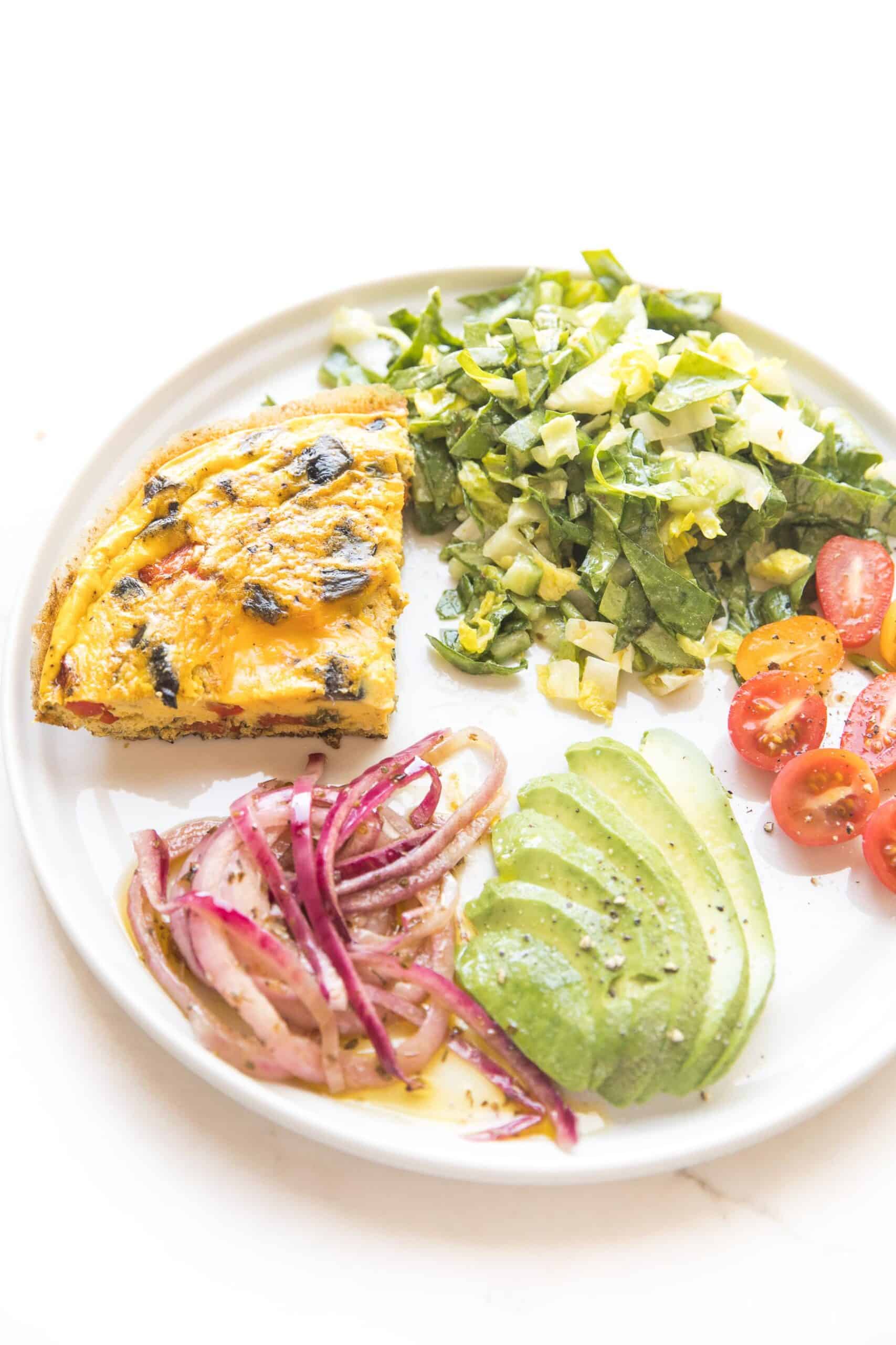 frittata on a white plate with marinated onions, avocado, tomatoes, green salad