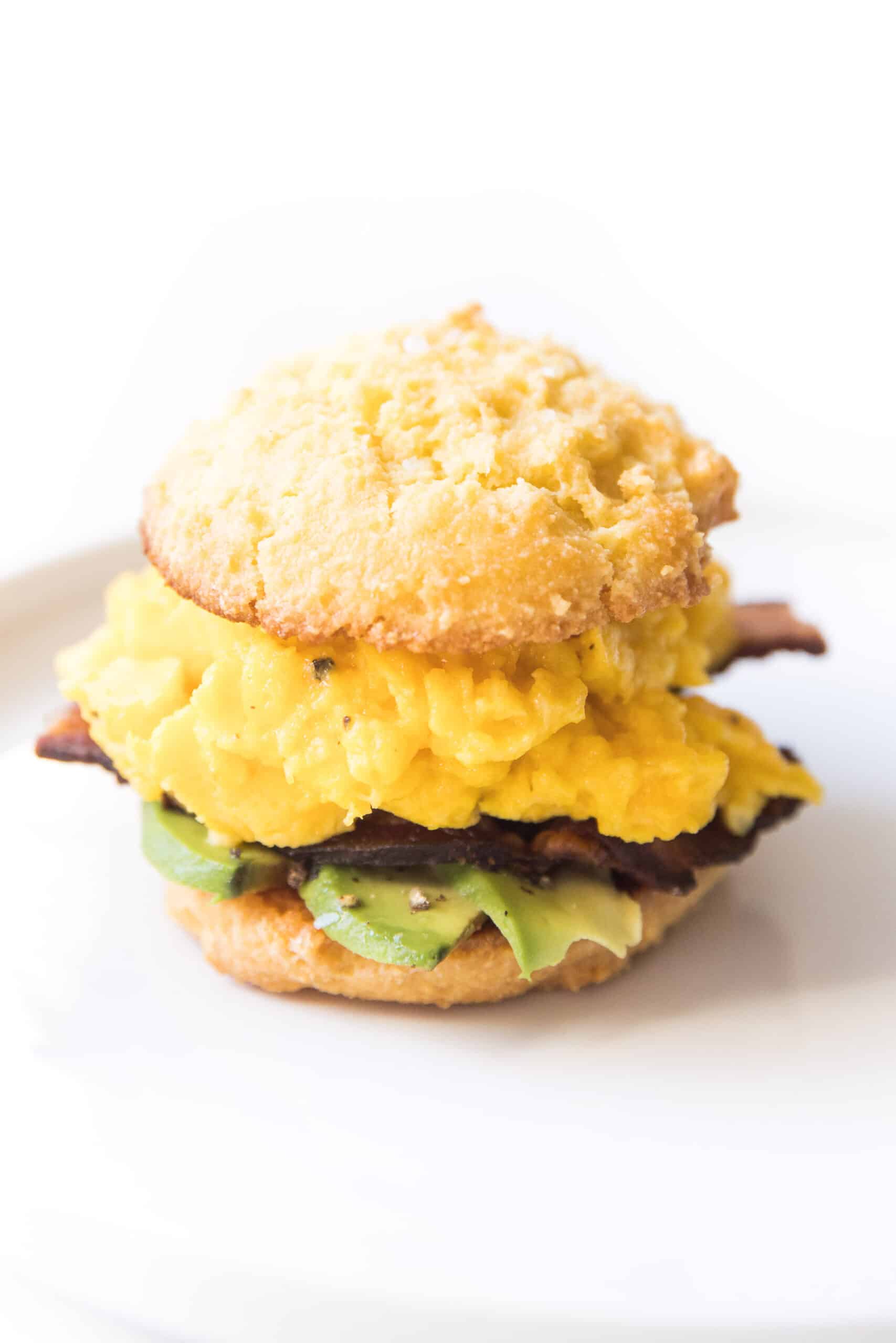 keto biscuit breakfast sandwich with egg, bacon and avocado on a white plate and background