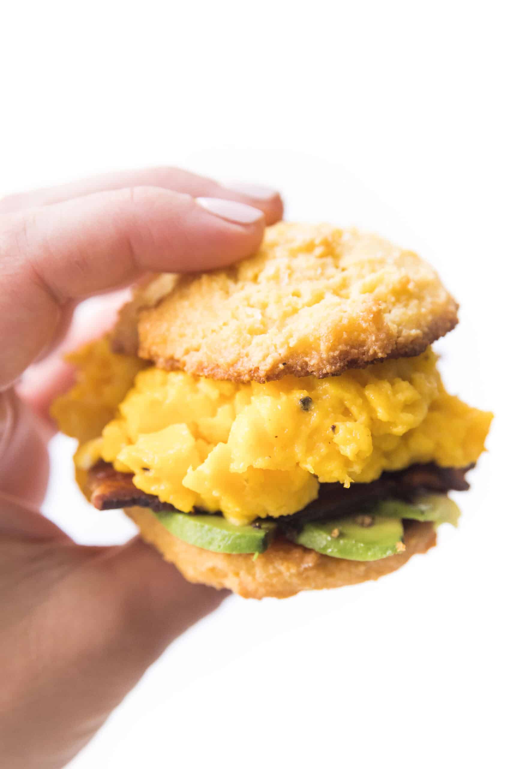 hand holding a keto biscuit breakfast sandwich with egg, bacon and avocado on a white plate and background