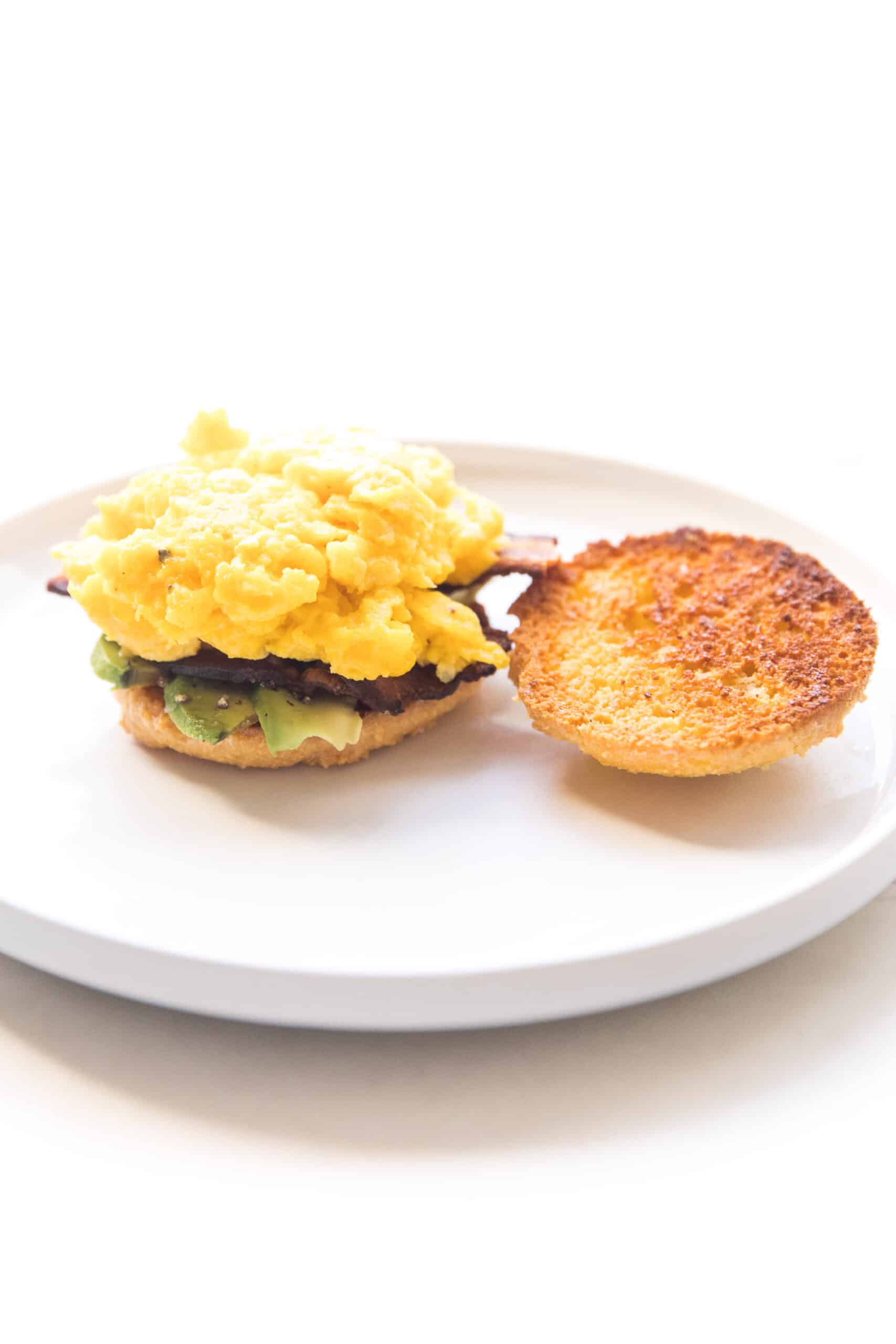 keto biscuit breakfast sandwich with egg, bacon and avocado on a white plate and background