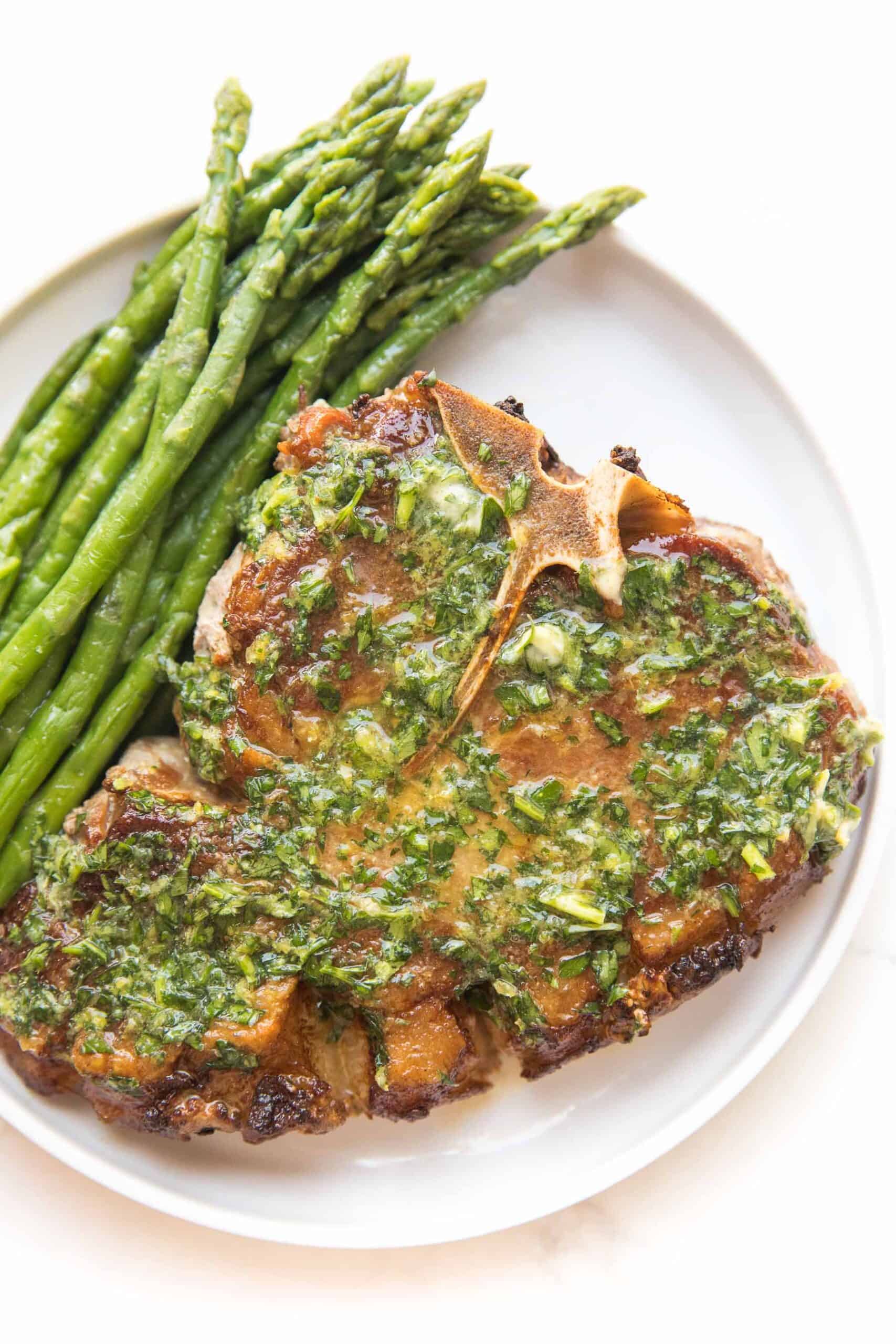 pork chop covered in melted herb butter with parsley and asparagus on a white plate and background