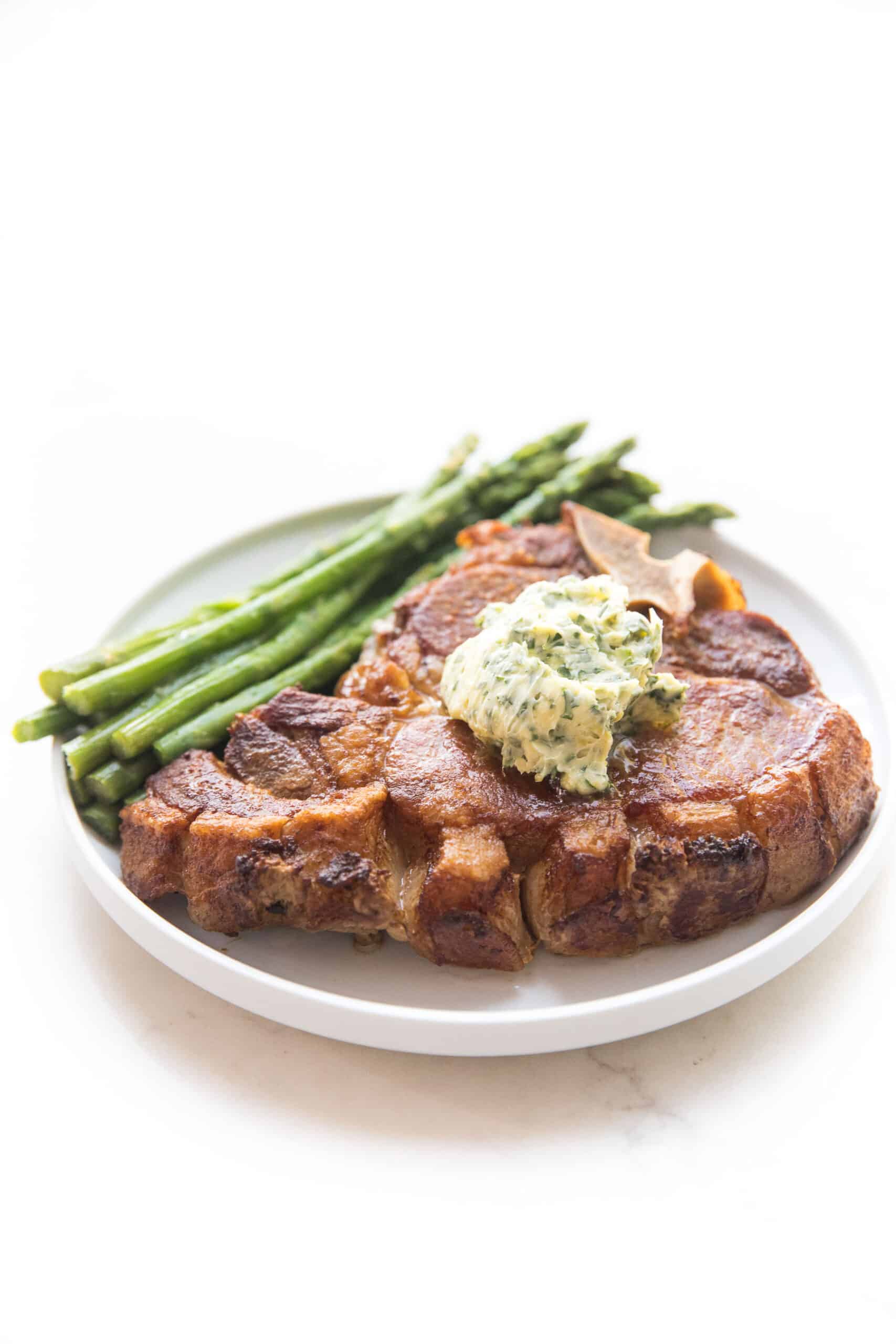 pork chop topped with herb butter with parsley and asparagus on a white plate and background