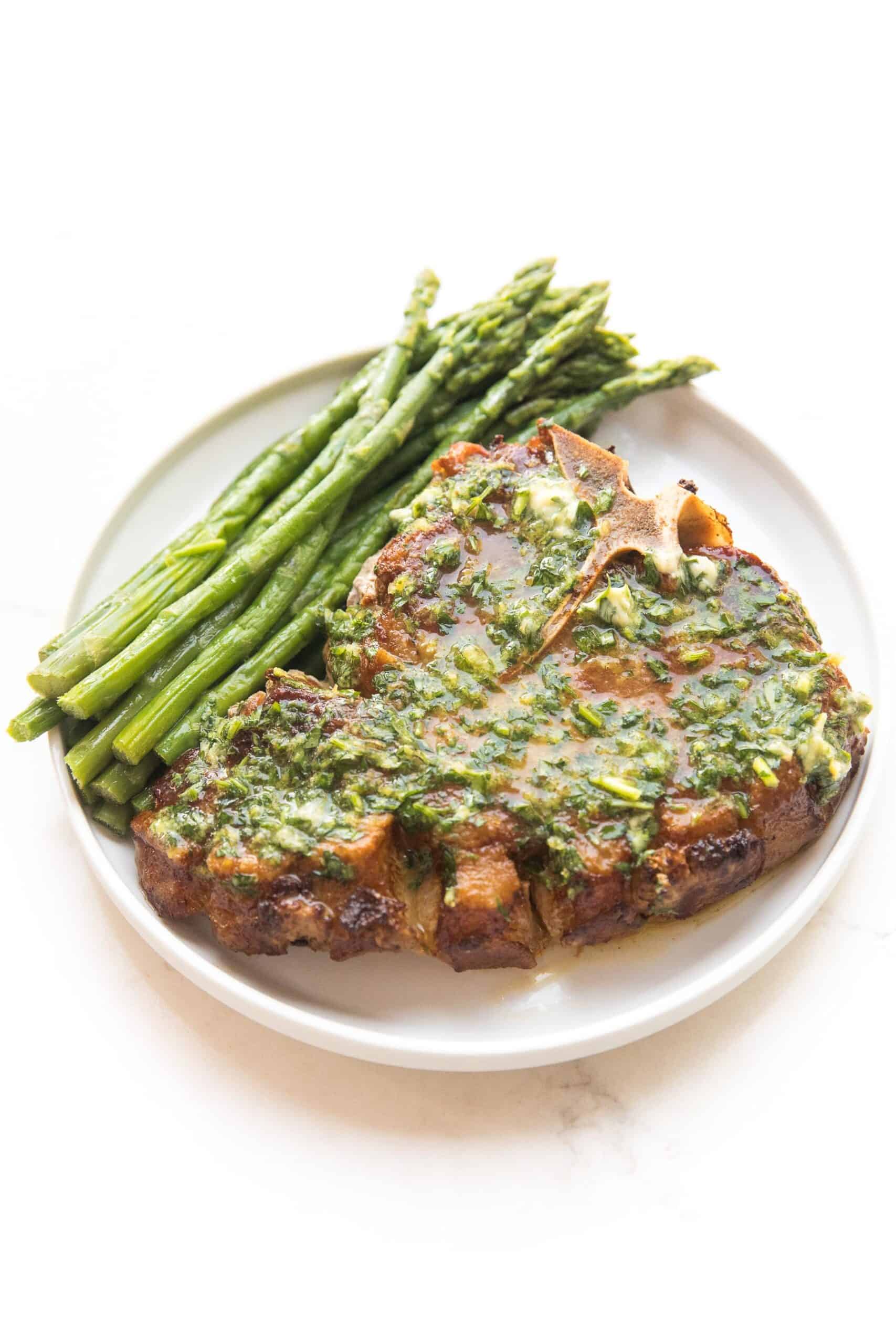 pork chop covered in melted herb butter with parsley and asparagus on a white plate and background