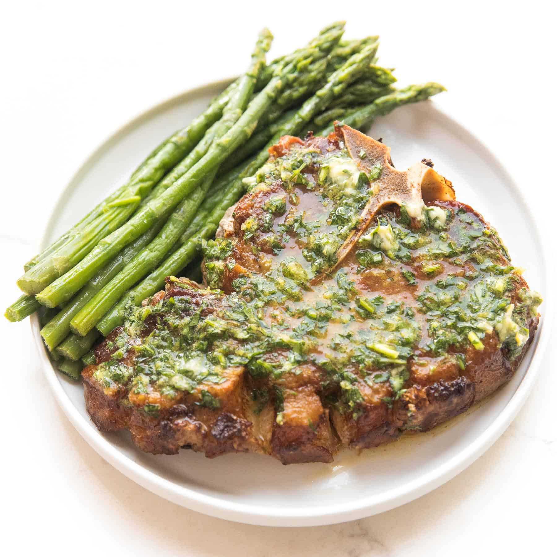 pork chop covered in melted herb butter with parsley and asparagus on a white plate and background