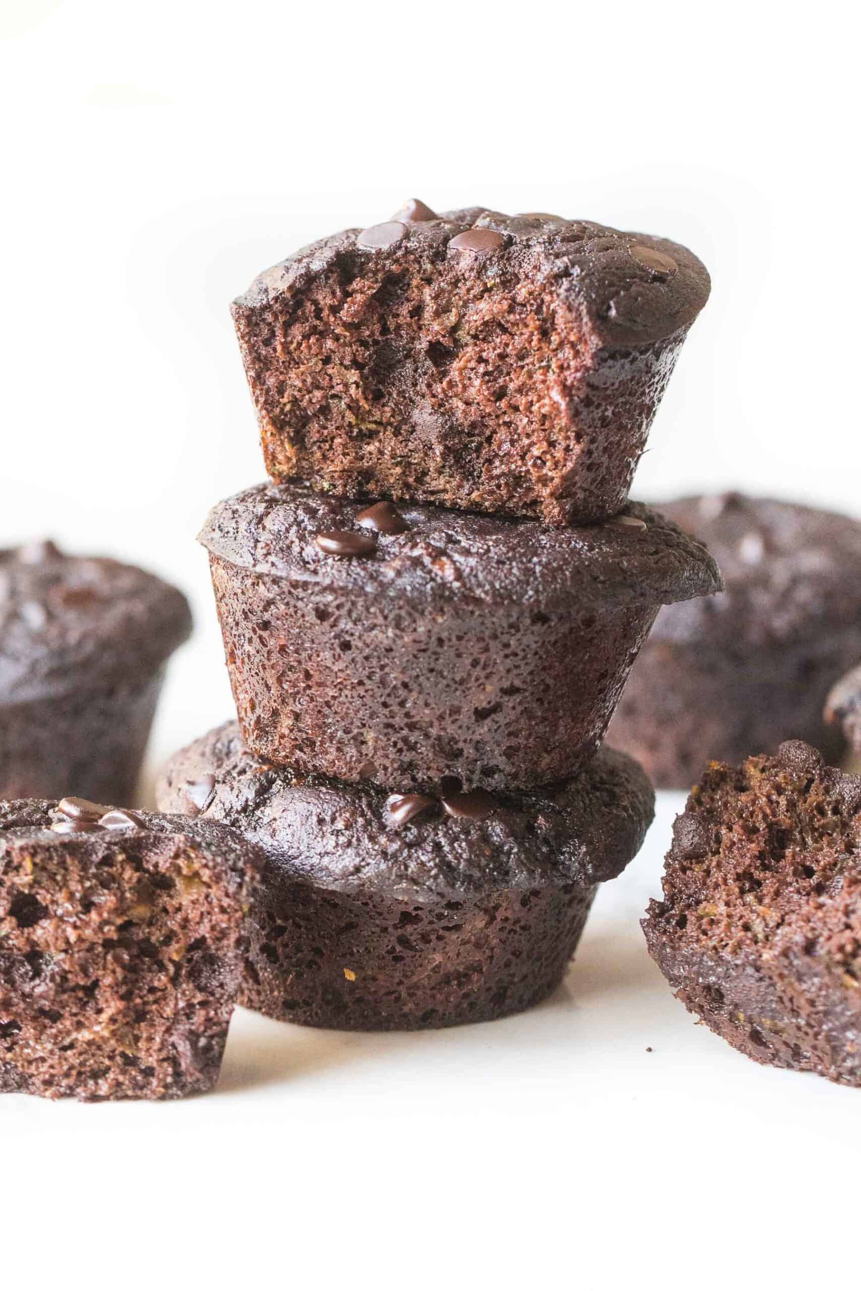 keto chocolate zucchini muffins stacked with a bite taken out on a white background
