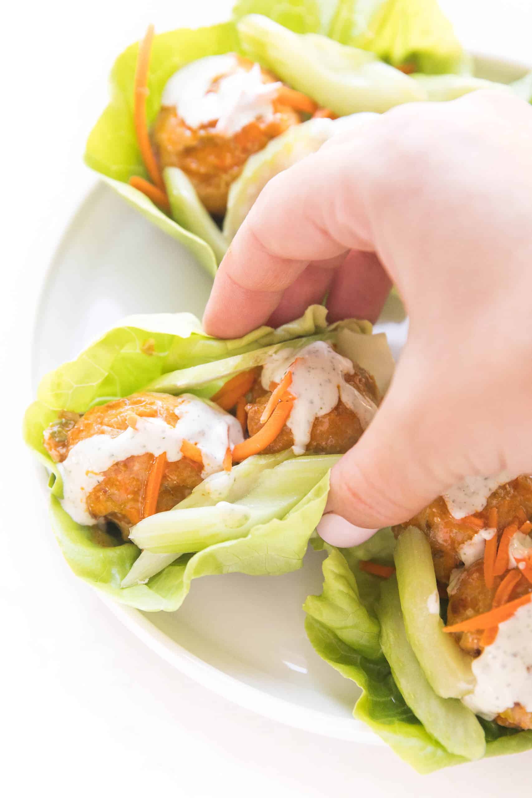 hand grabbing picking up keto buffalo turkey meatballs on lettuce wraps topped with ranch dressing on a white plate and background