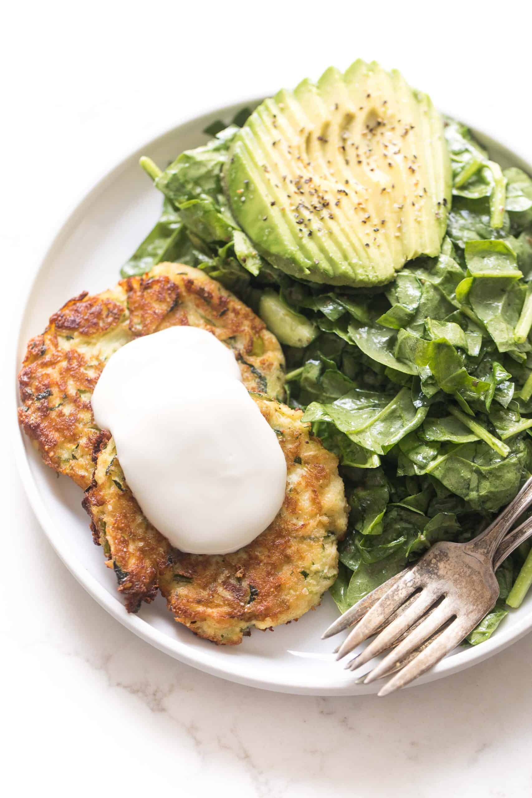 zucchini fritters topped with sour cream on a white plate and background with spinach salad + avocado