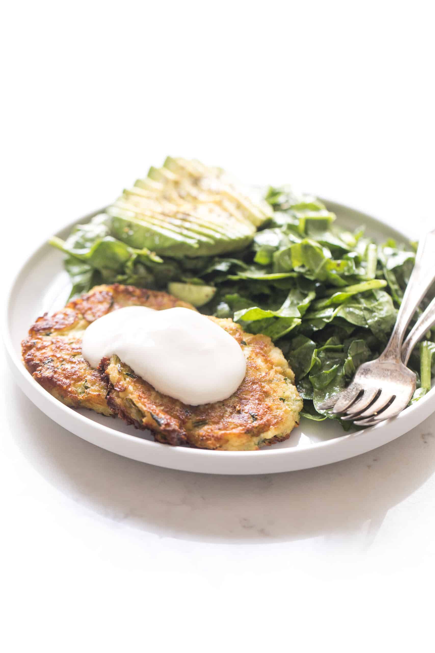 zucchini fritters topped with sour cream on a white plate and background with spinach salad + avocado