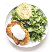 zucchini fritters topped with sour cream on a white plate and background with spinach salad + avocado
