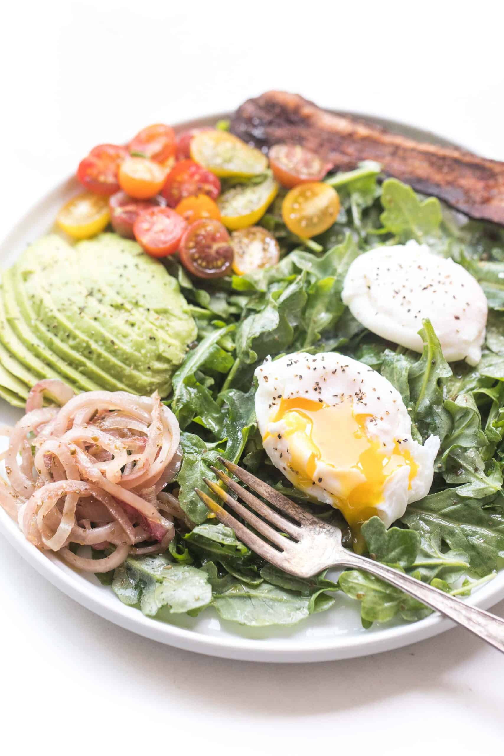 breakfast salad with baby arugula, poached egg, onions, avocado, tomato and bacon on a white plate and background