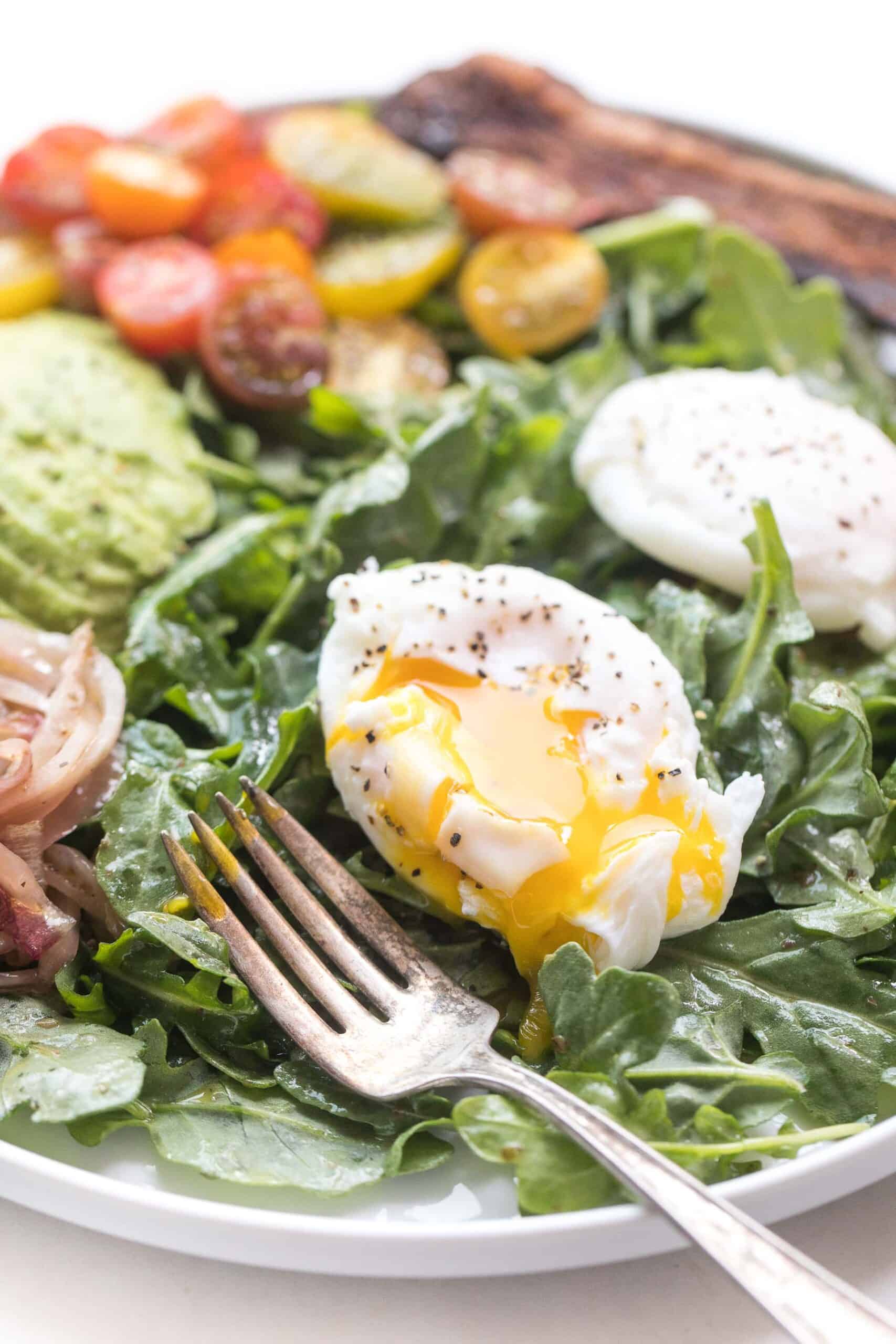 breakfast salad with baby arugula, poached egg, onions, avocado, tomato and bacon on a white plate and background