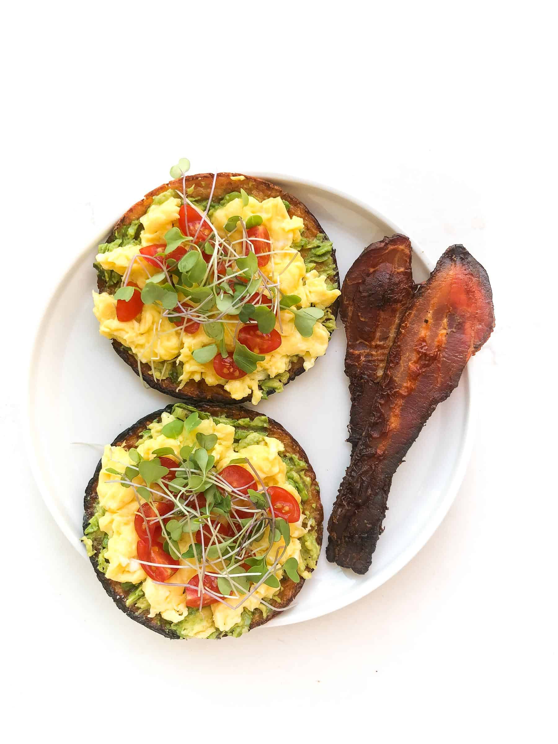 keto avocado toast topped with egg, tomatoes, microgreens and bacon on a white plate and background