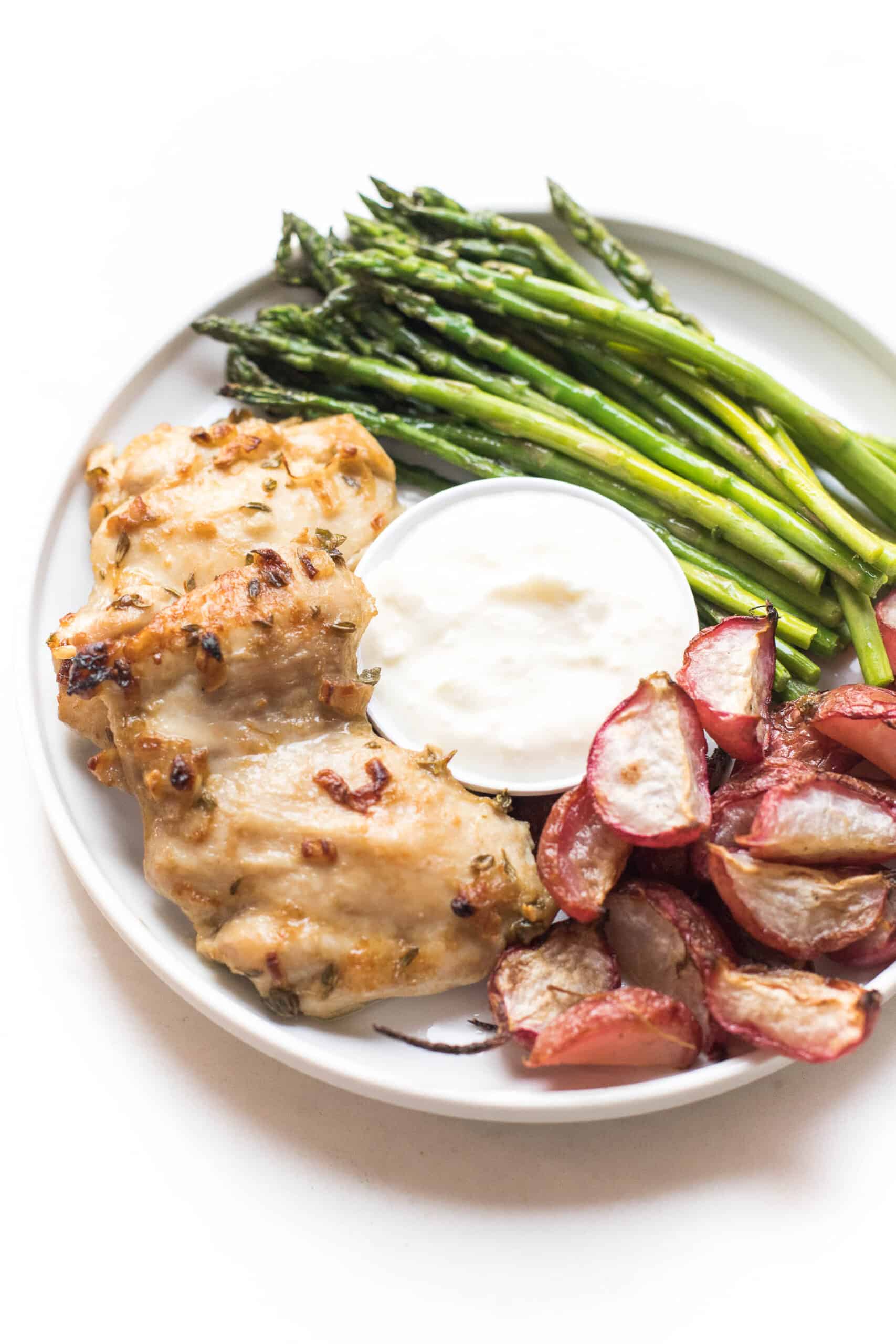 Dijon mustard chicken thighs with radishes, asparagus and yogurt feta dip on a white plate and background