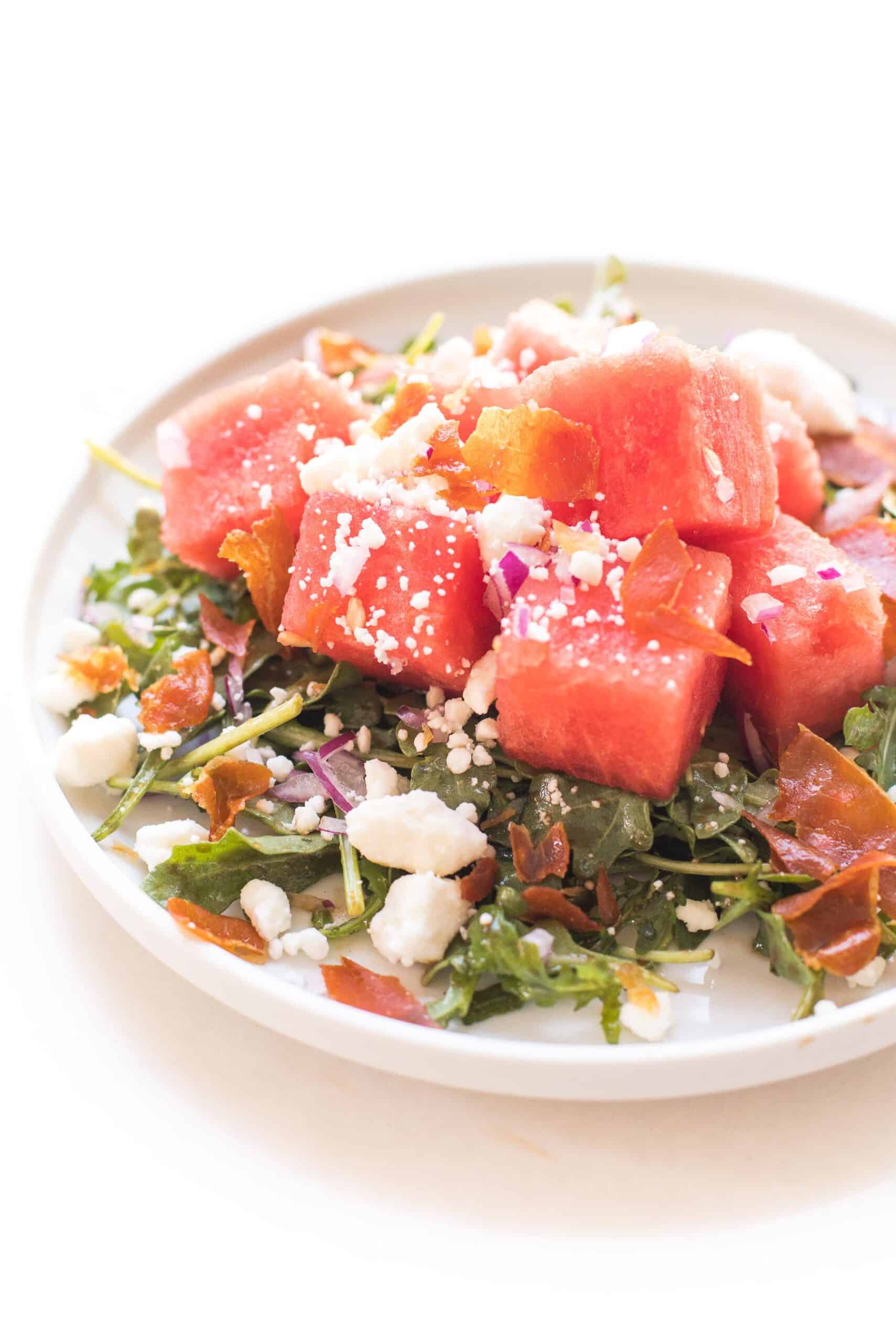 WATERMELON FETA SALAD WITH ARUGULA, CRISPY PROSCIUTTO AND RED ONION ON A WHITE PLATE