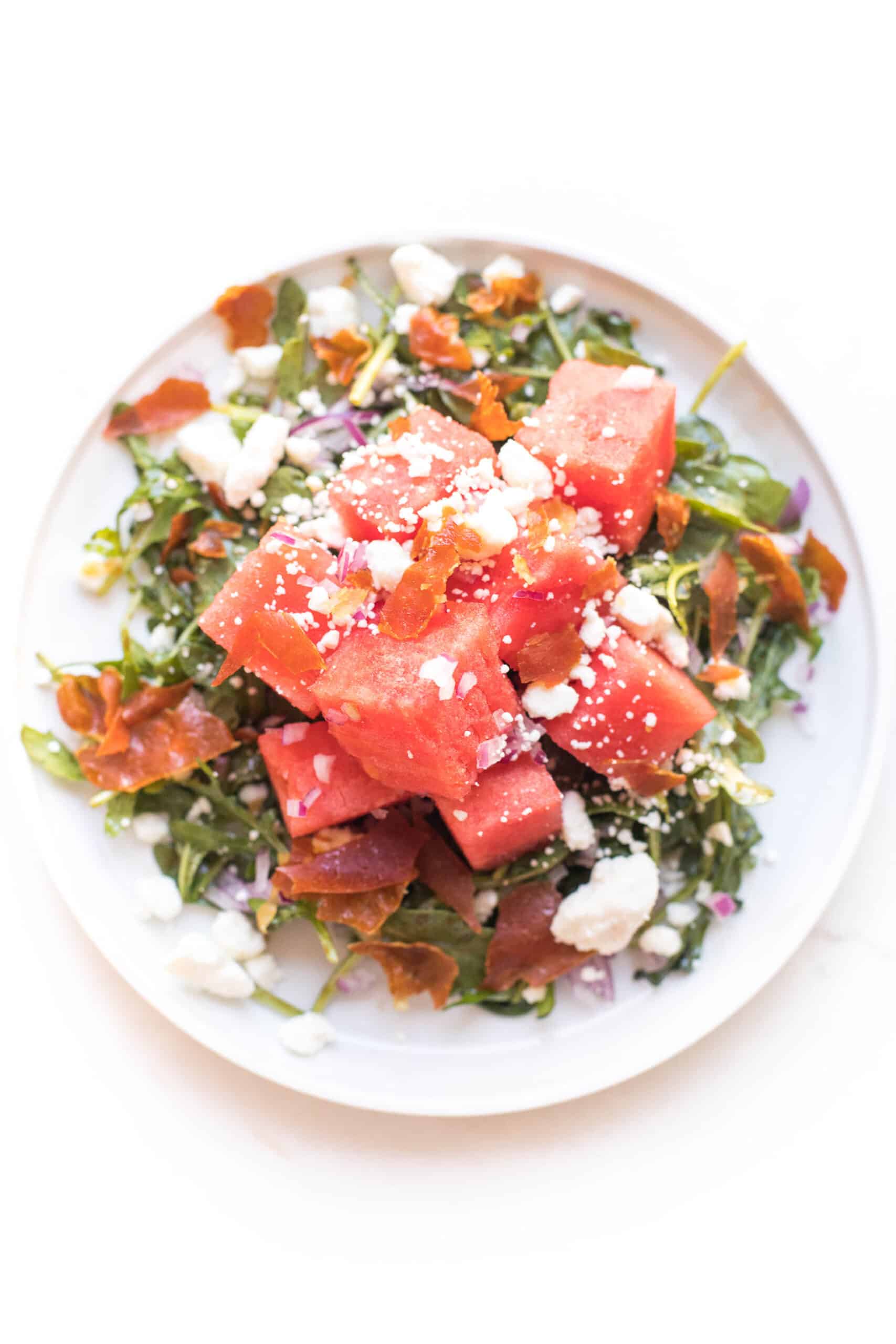 WATERMELON FETA SALAD WITH ARUGULA, CRISPY PROSCIUTTO AND RED ONION ON A WHITE PLATE