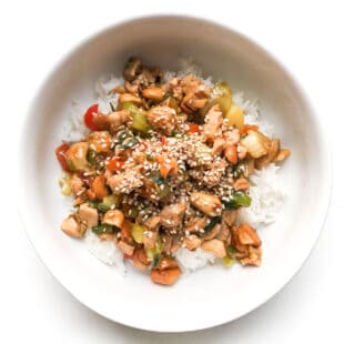 CHICKEN AND CELERY STIR FRY IN A WHITE BOWL TOPPED WITH SESAME SEEDS OVER CAULIFLOWER RICE IN A WHITE BOWL AND BACKGROUND
