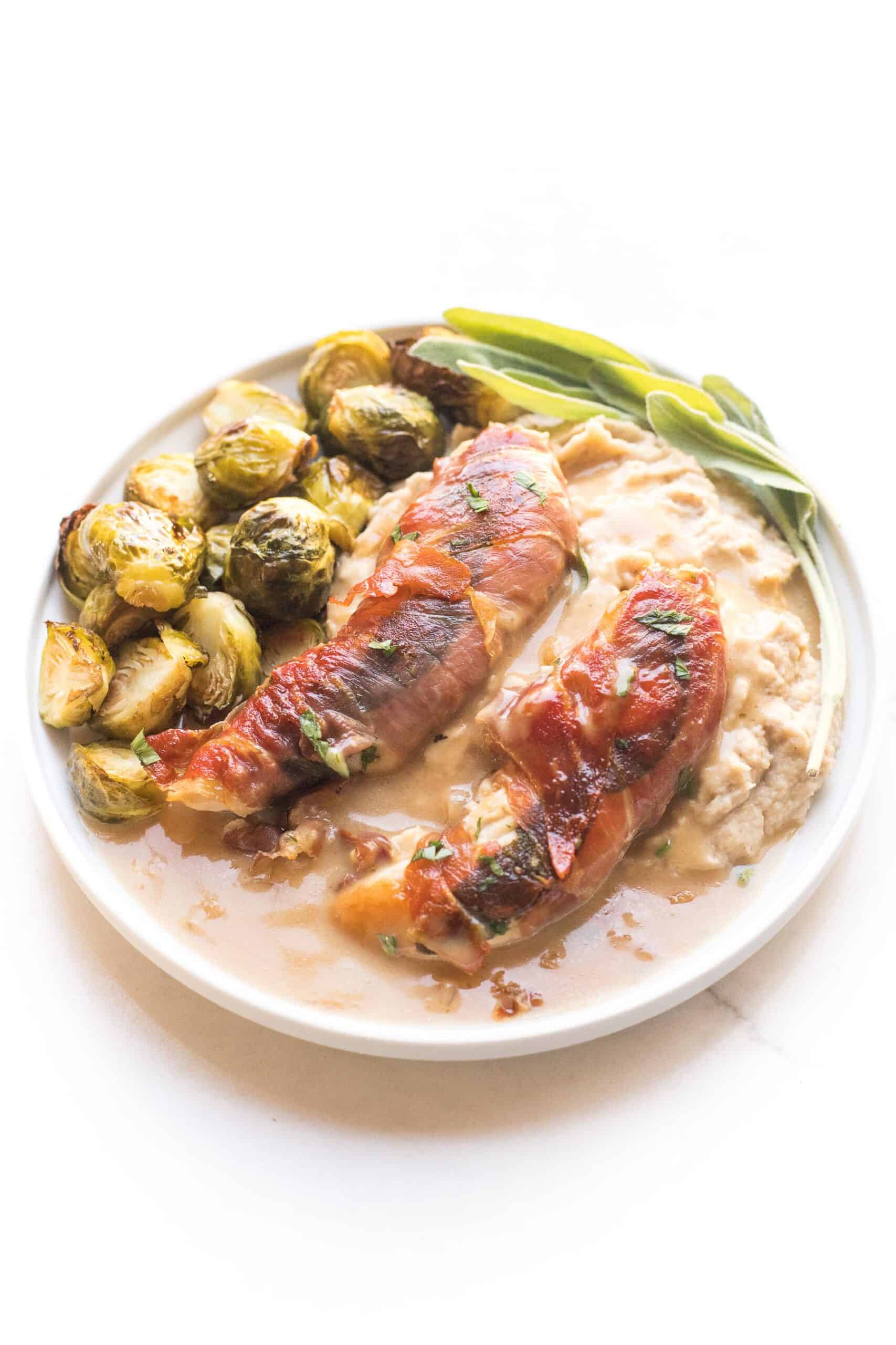 keto + whole30 prosciutto wrapped chicken with creamy apple cider gravy over mashed cauliflower with roasted brussels sprouts on a white plate and background
