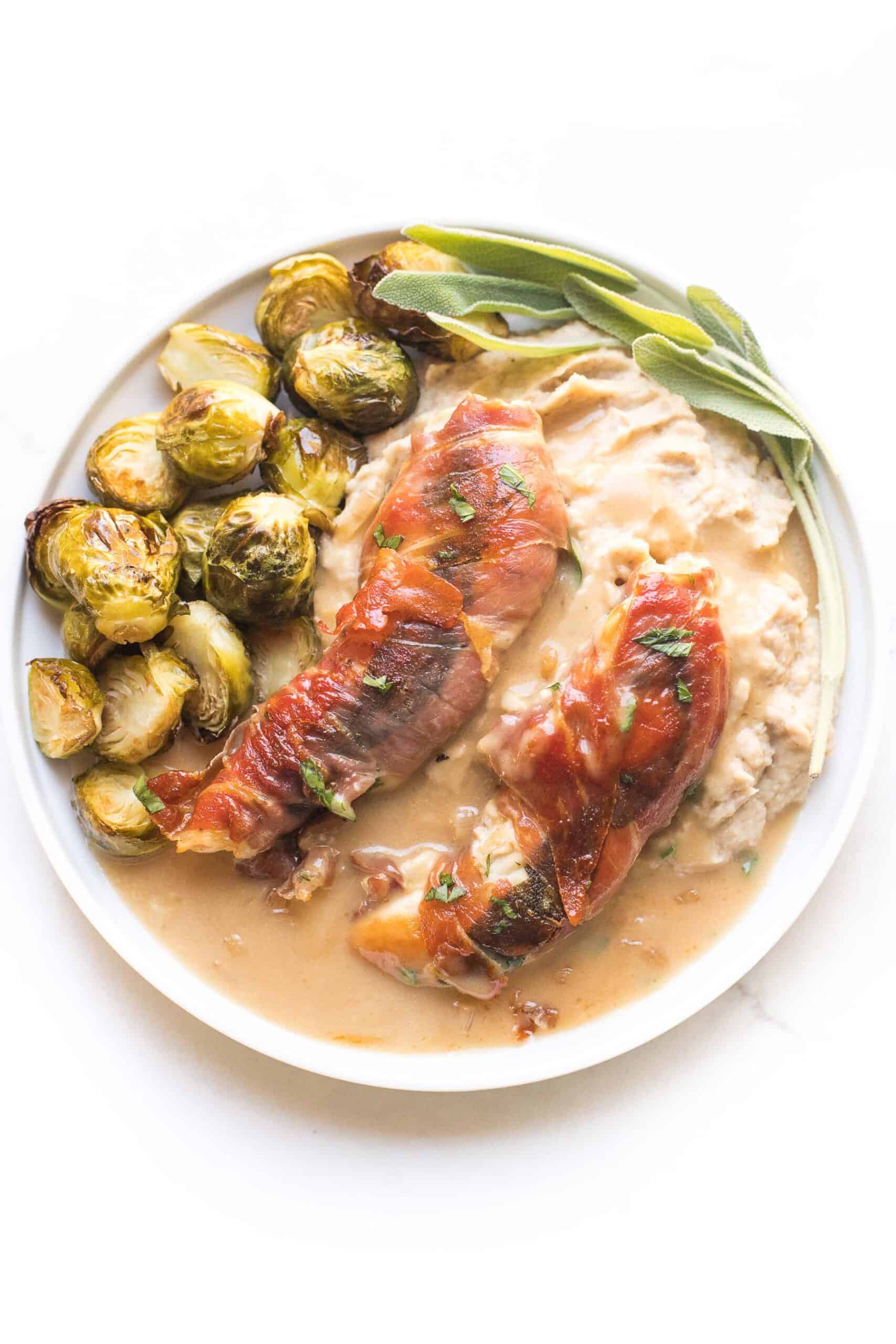 keto + whole30 prosciutto wrapped chicken with creamy apple cider gravy over mashed cauliflower with roasted brussels sprouts on a white plate and background