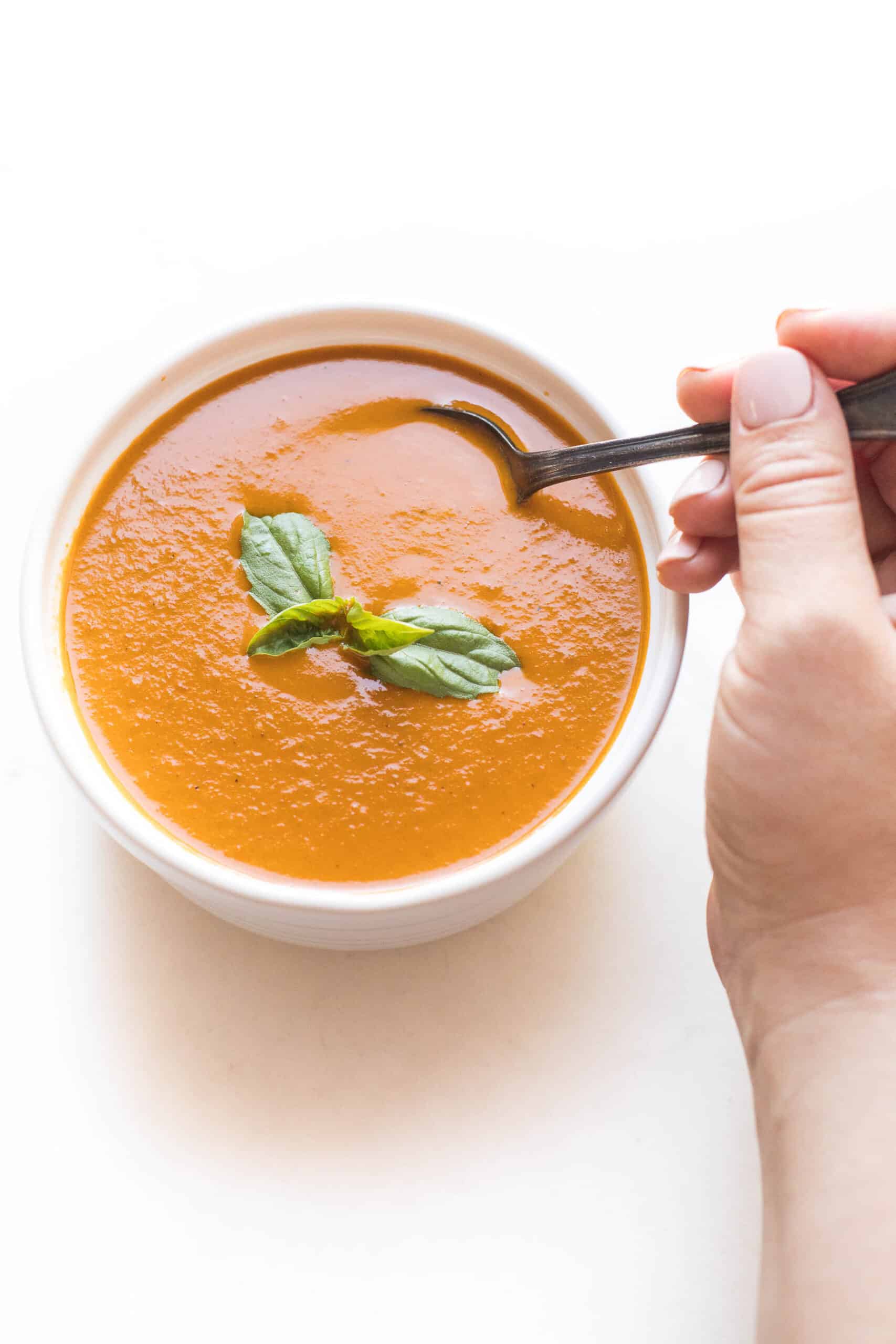 hand spooning roasted tomato basil soup in a white bowl on a white background