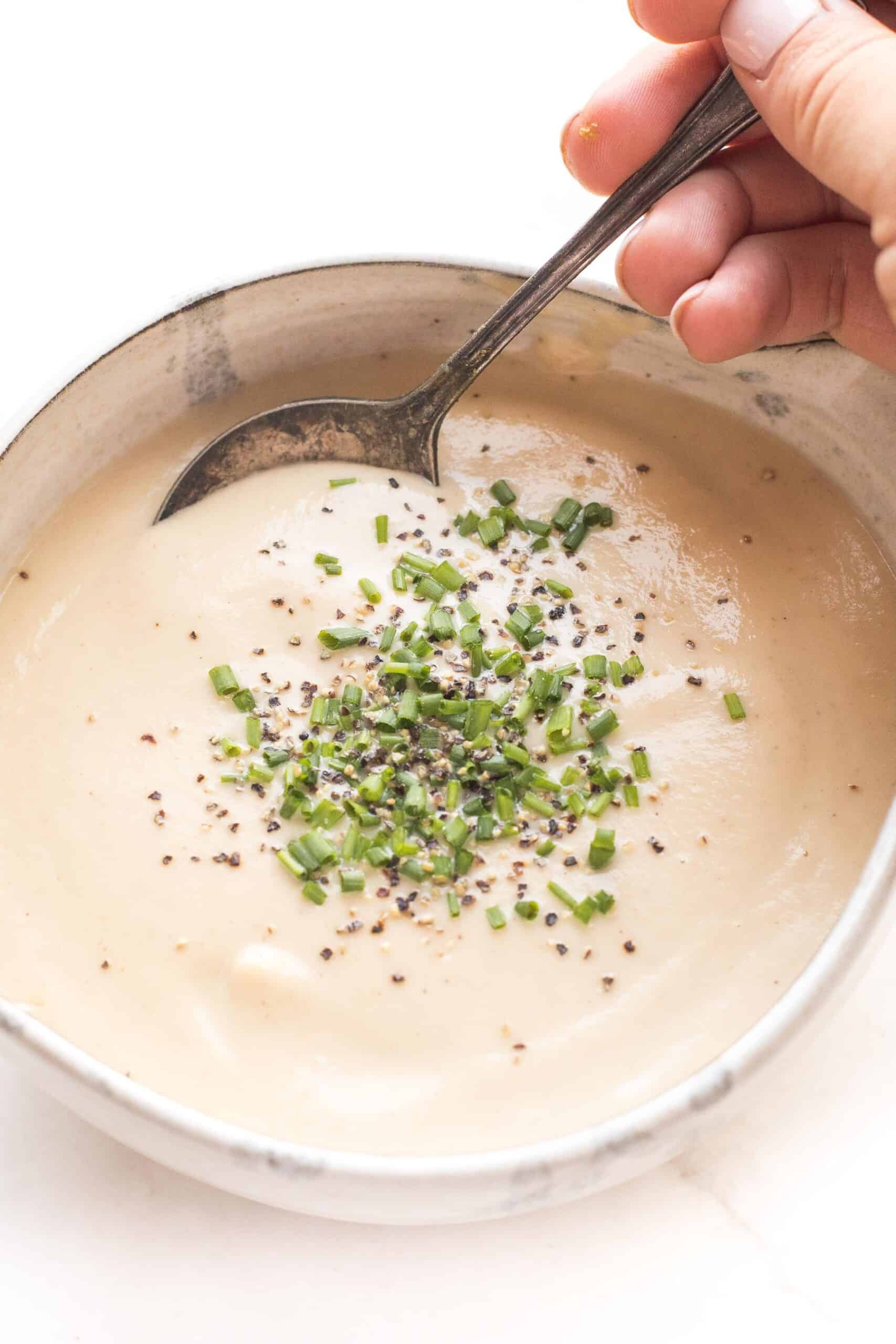 hand holding a spoon scooping out keto roasted cauliflower soup in a pottery bowl topped with chives and black pepper