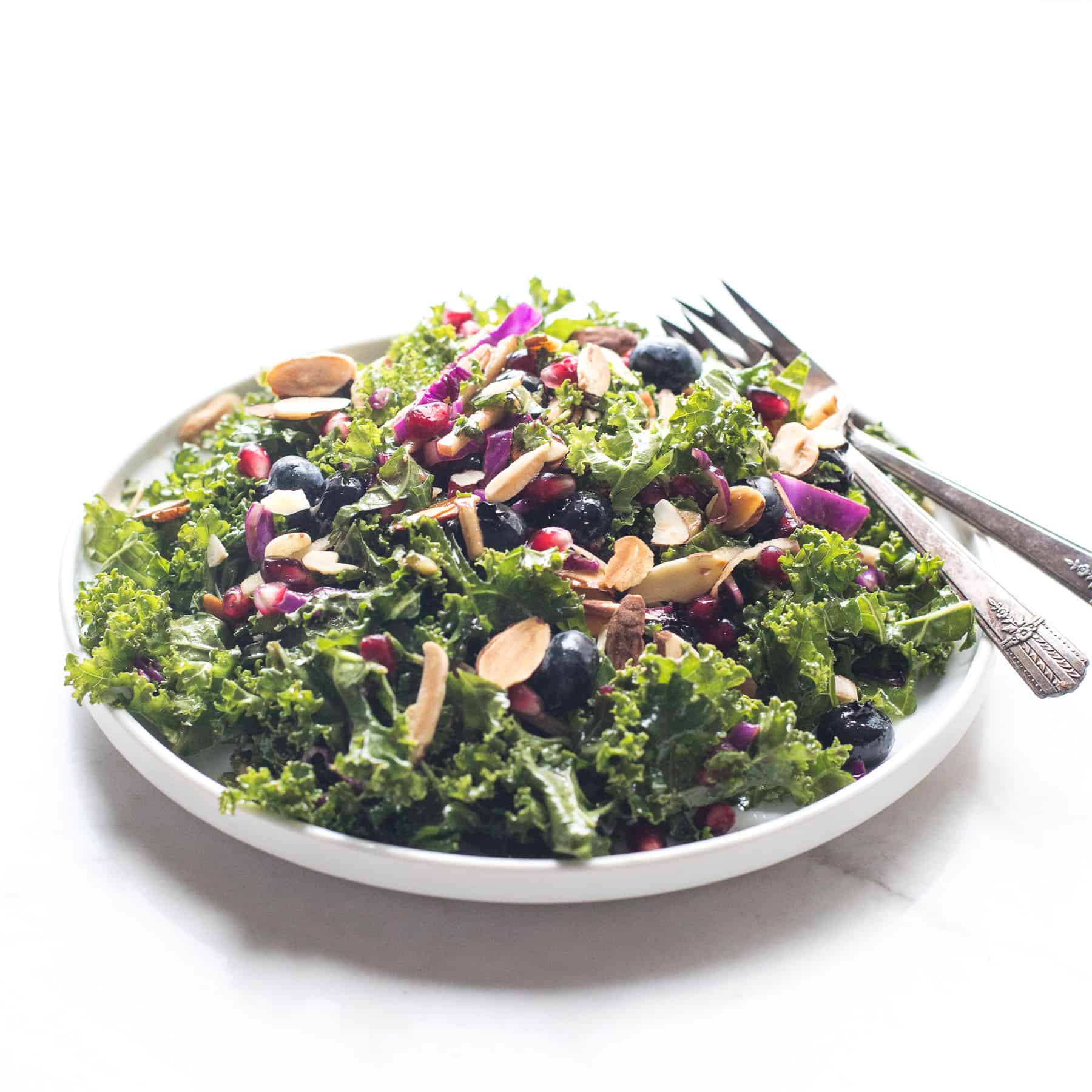 Whole30 + Keto Pomegranate + Blueberry Winter Kale Salad with almonds, green onions, red cabbage and apples on a white plate and background