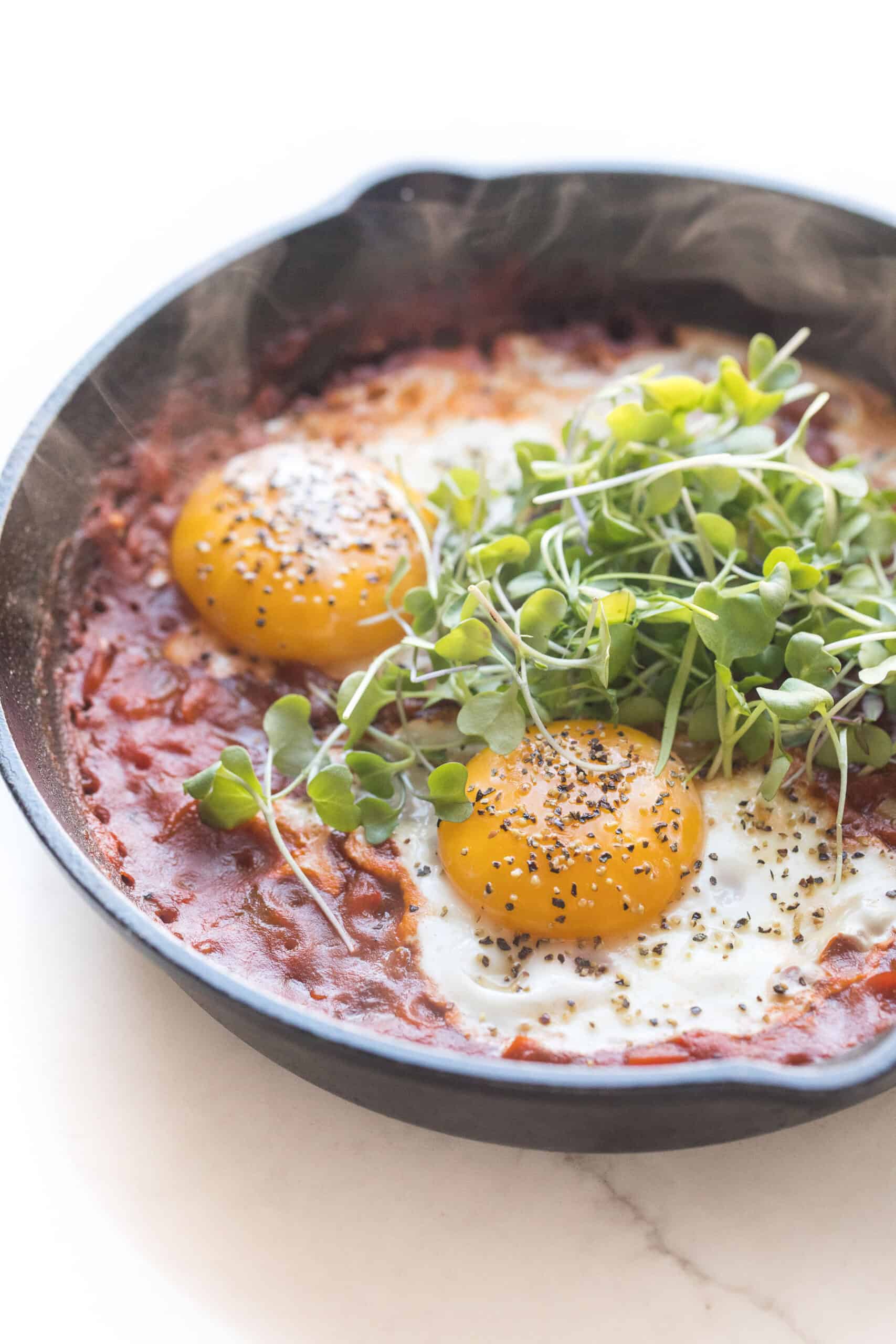 KETO SHAKSHUKA EGGS IN TOMATO SAUCE IN A CAST IRON SKILLET ON A WHITE BACKGROUND TOPPED WITH GREENS