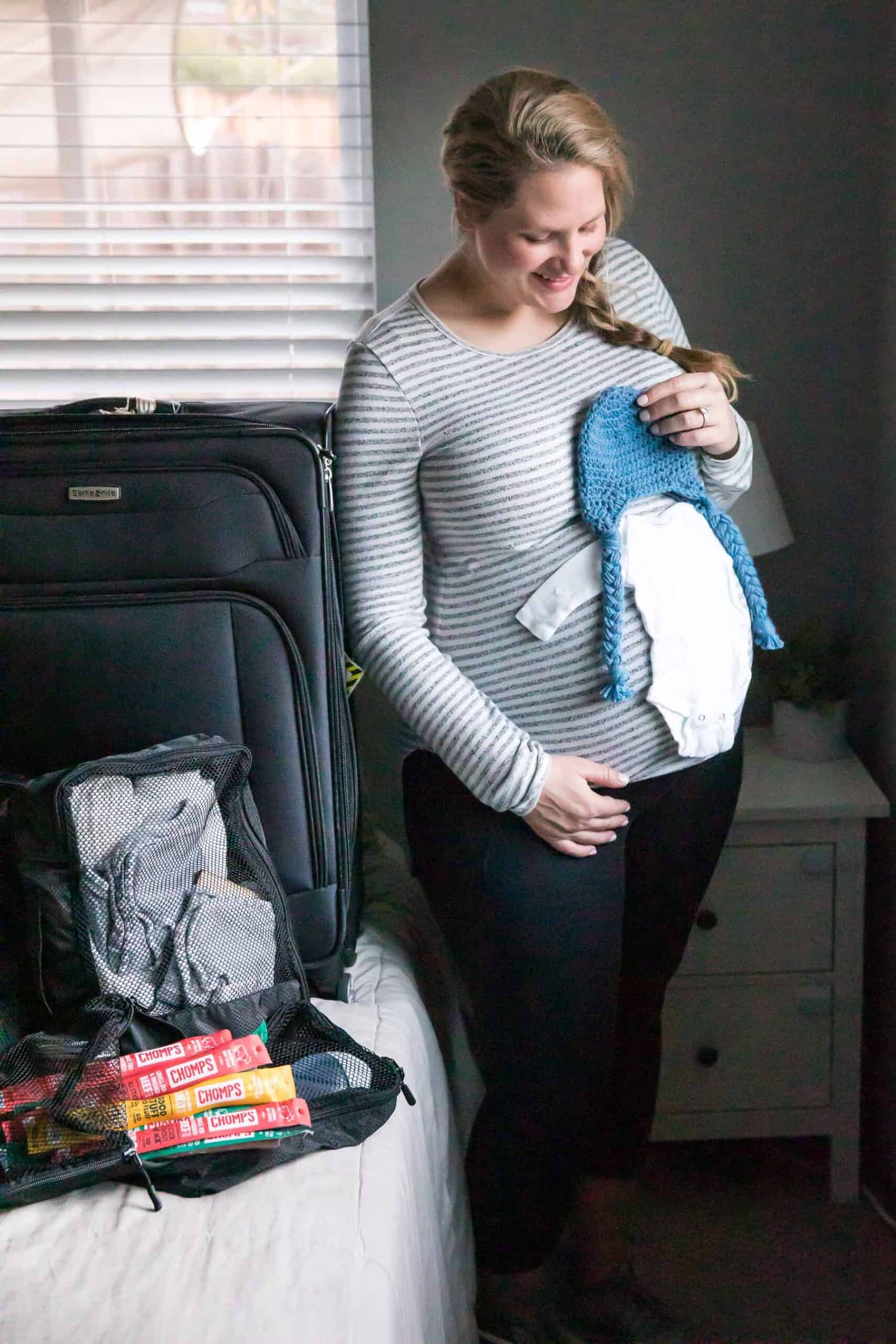 pregnant girl standing next to hospital bag holding up baby clothes