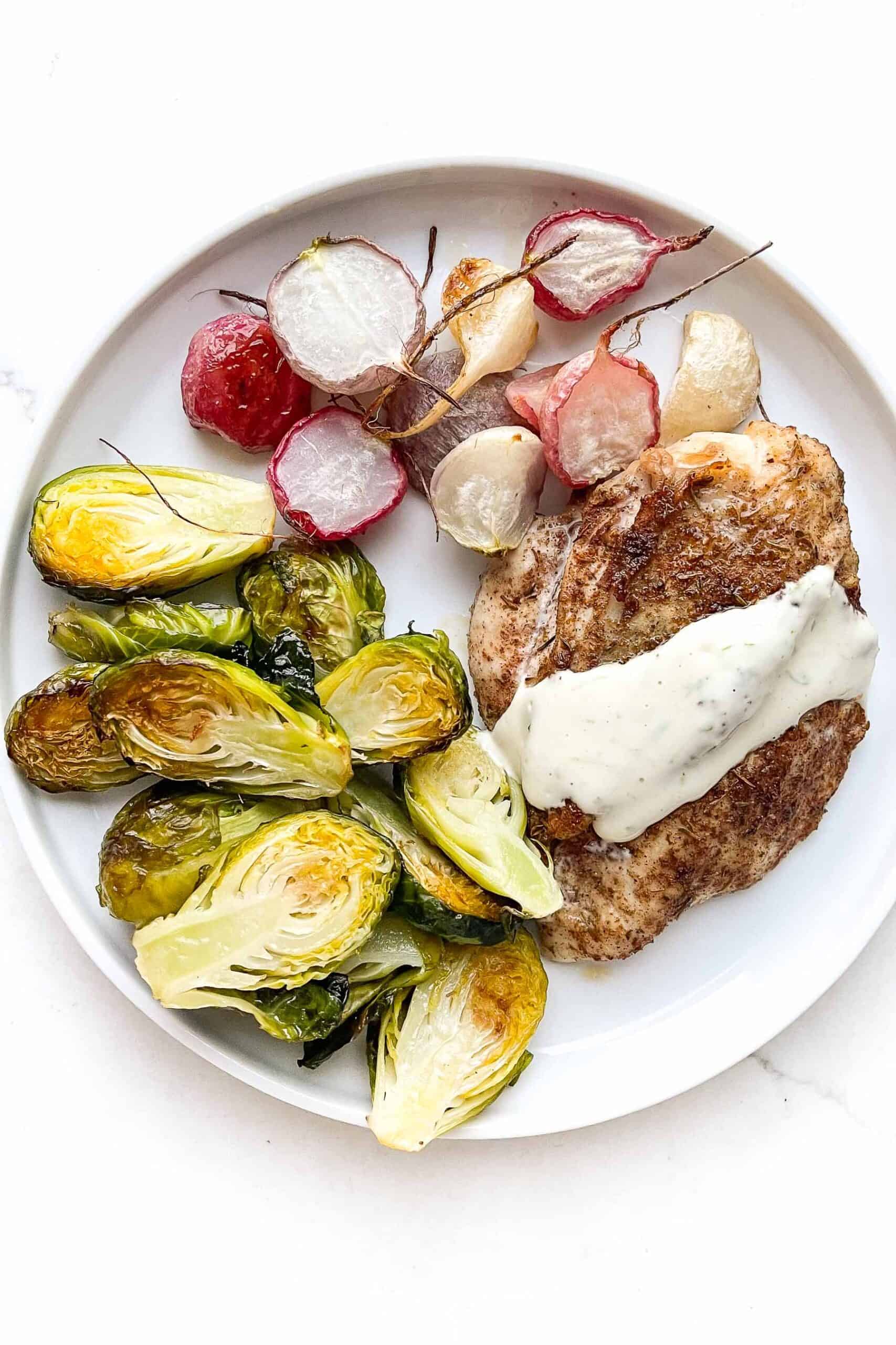 Jerk seasoned chicken breast topped with garlic lime aioli, roasted brussels sprouts + roasted radishes on a white plate and background