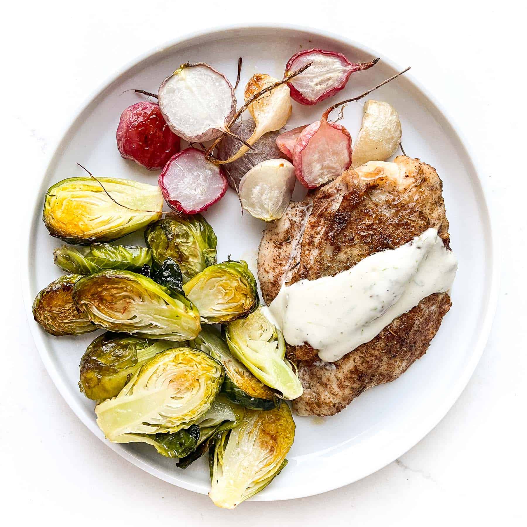 Jerk seasoned chicken breast topped with garlic lime aioli, roasted brussels sprouts + roasted radishes on a white plate and background