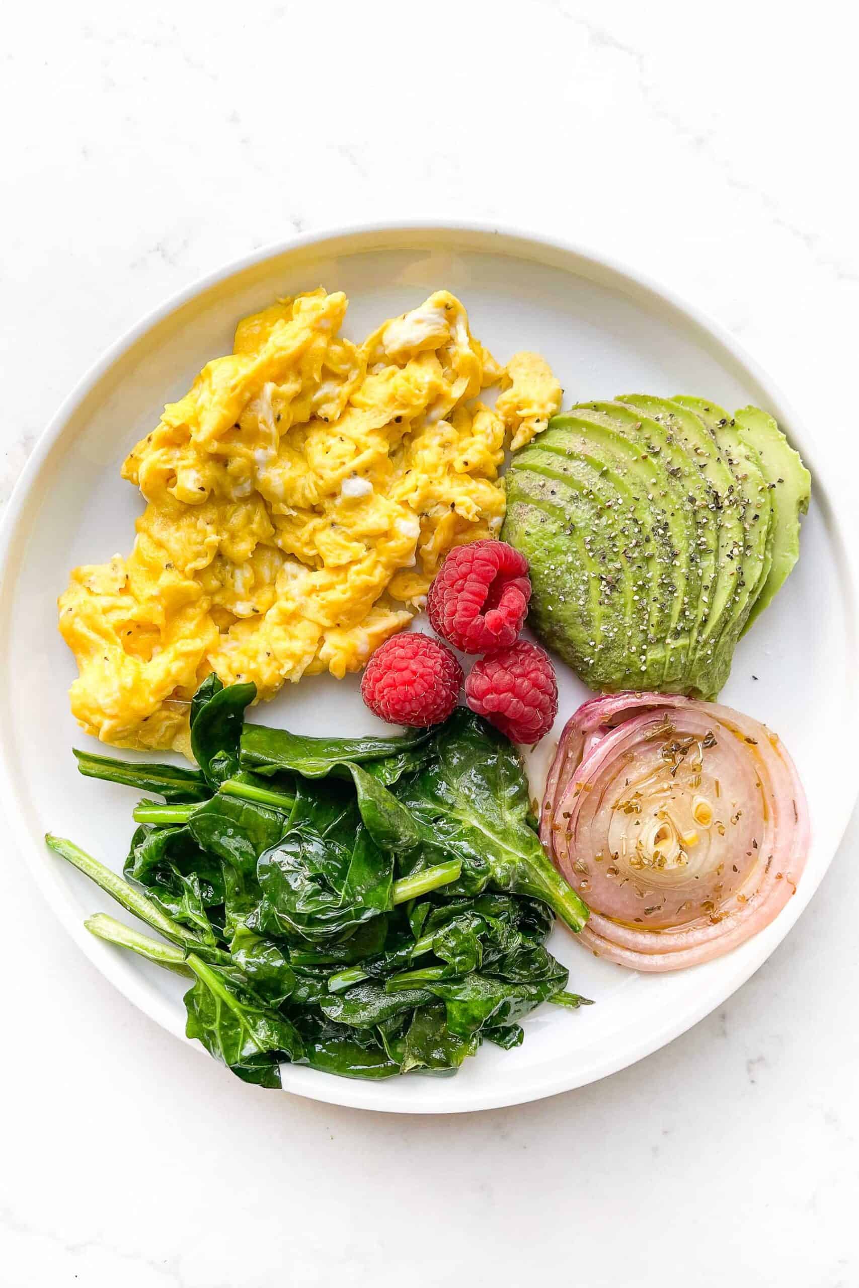 scrambled eggs, sauteed spinach, avocado, red onion and raspberries on a white plate and background