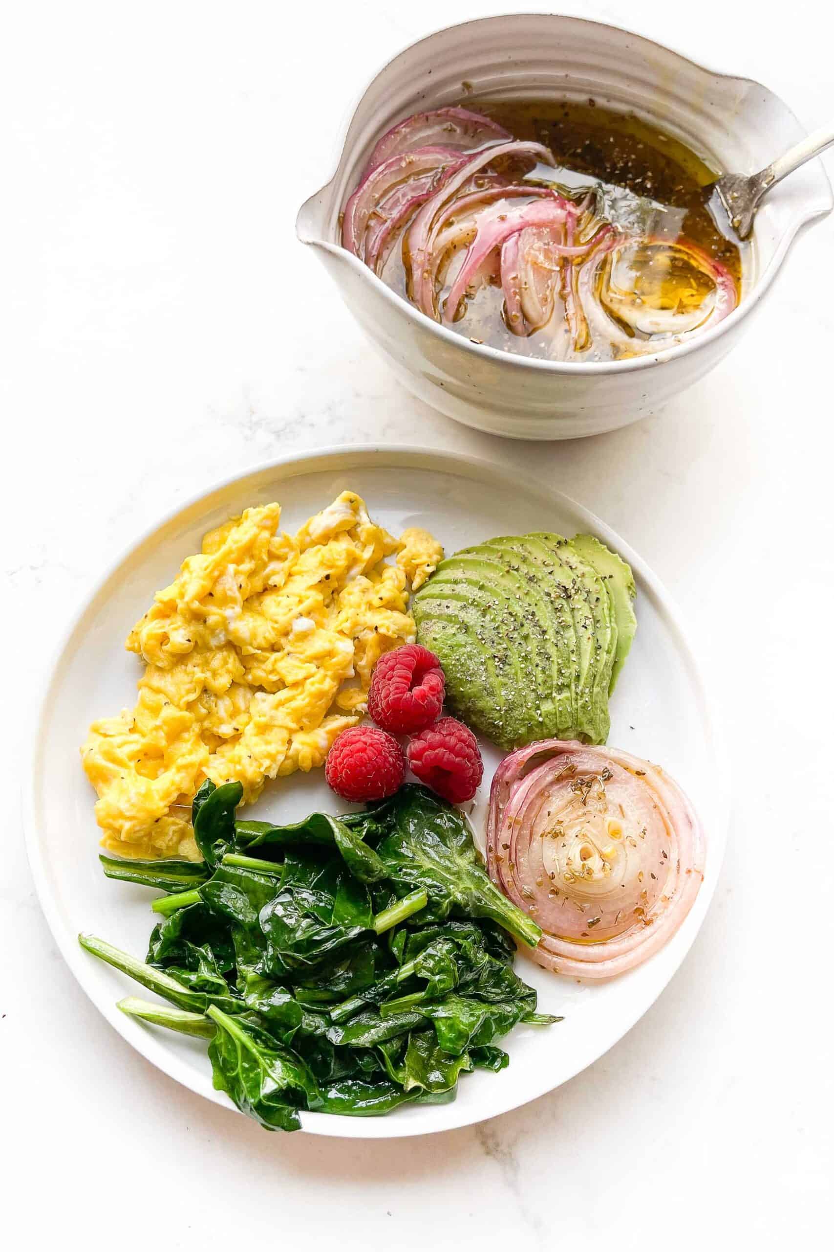 scrambled eggs, sauteed spinach, avocado, red onion and raspberries on a white plate and background