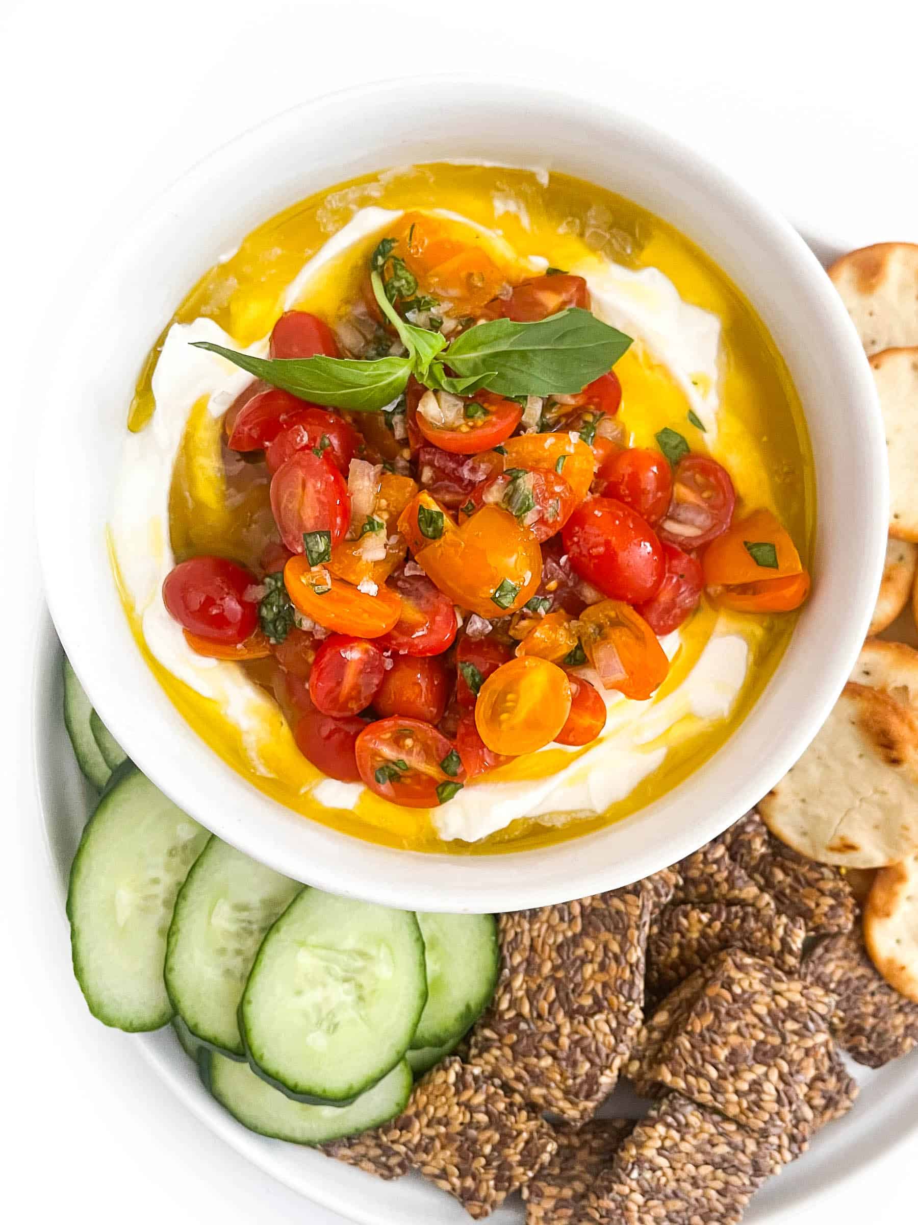 Whipped ricotta dip in a white bowl topped with grape tomatoes, basil and olive oil with cucumbers and crackers