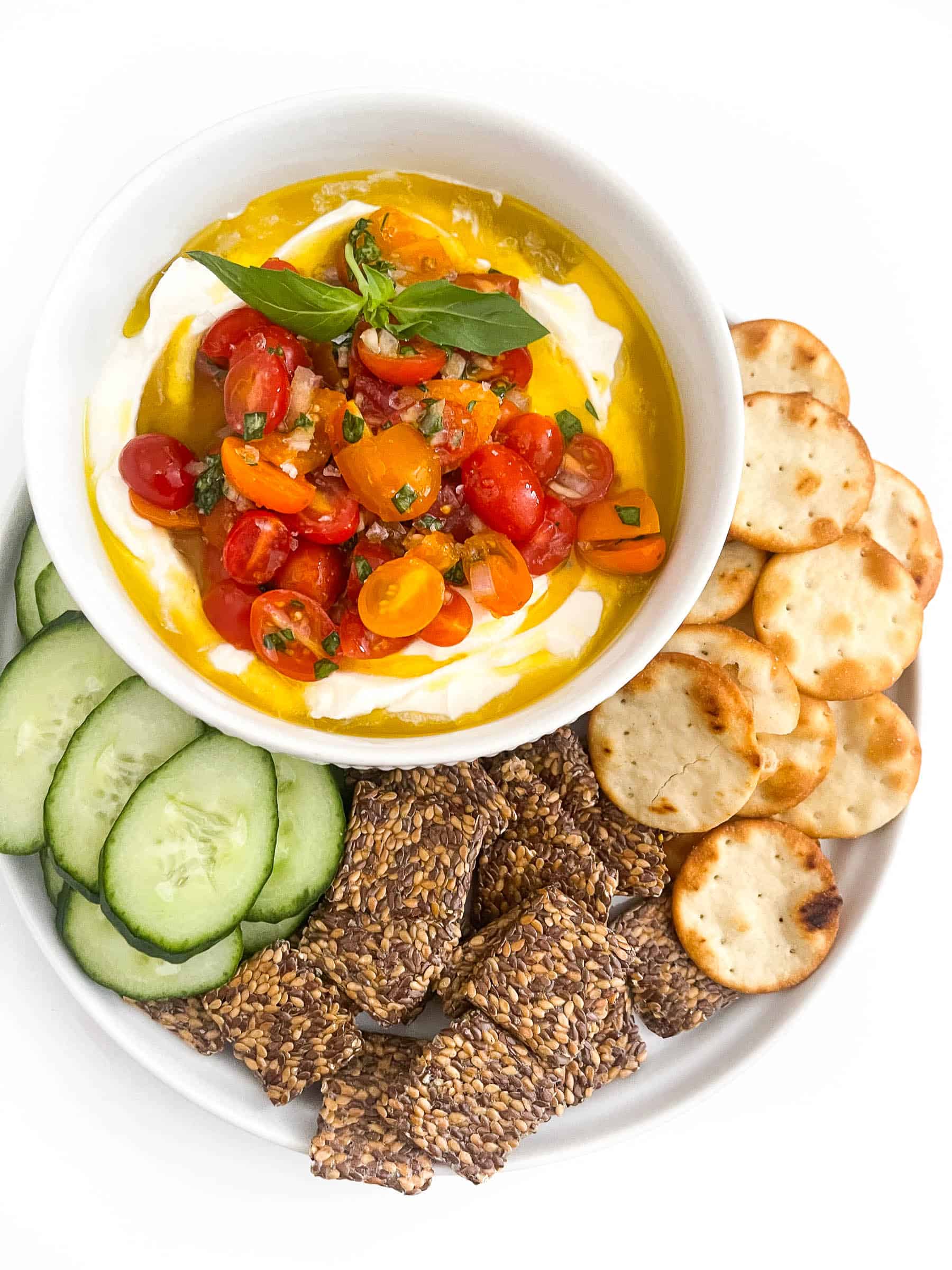 Whipped ricotta dip in a white bowl topped with grape tomatoes, basil and olive oil with cucumbers and crackers