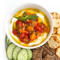 Whipped ricotta dip in a white bowl topped with grape tomatoes, basil and olive oil with cucumbers and crackers