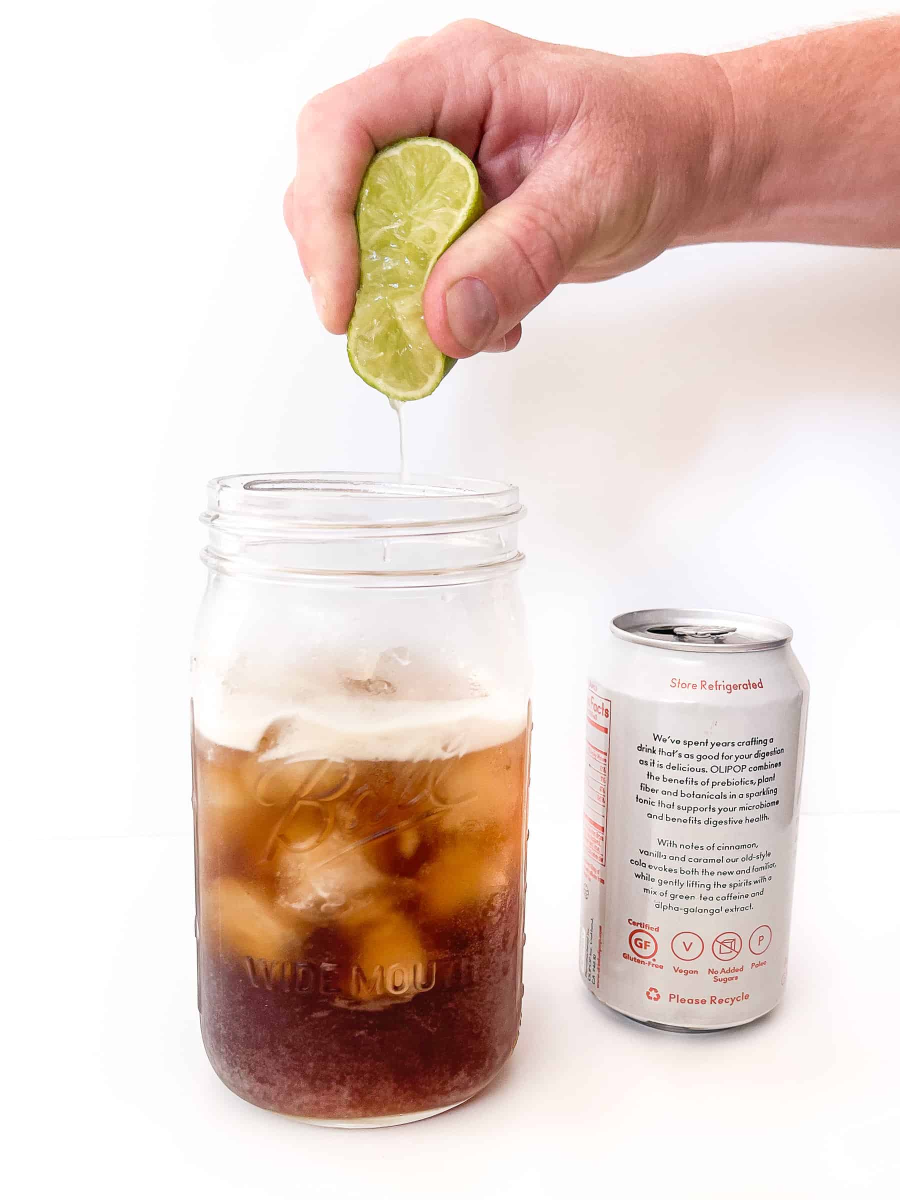 a hand squeezing half a lime into a dirty diet coke in a mason jar next to a can of olipop soda 