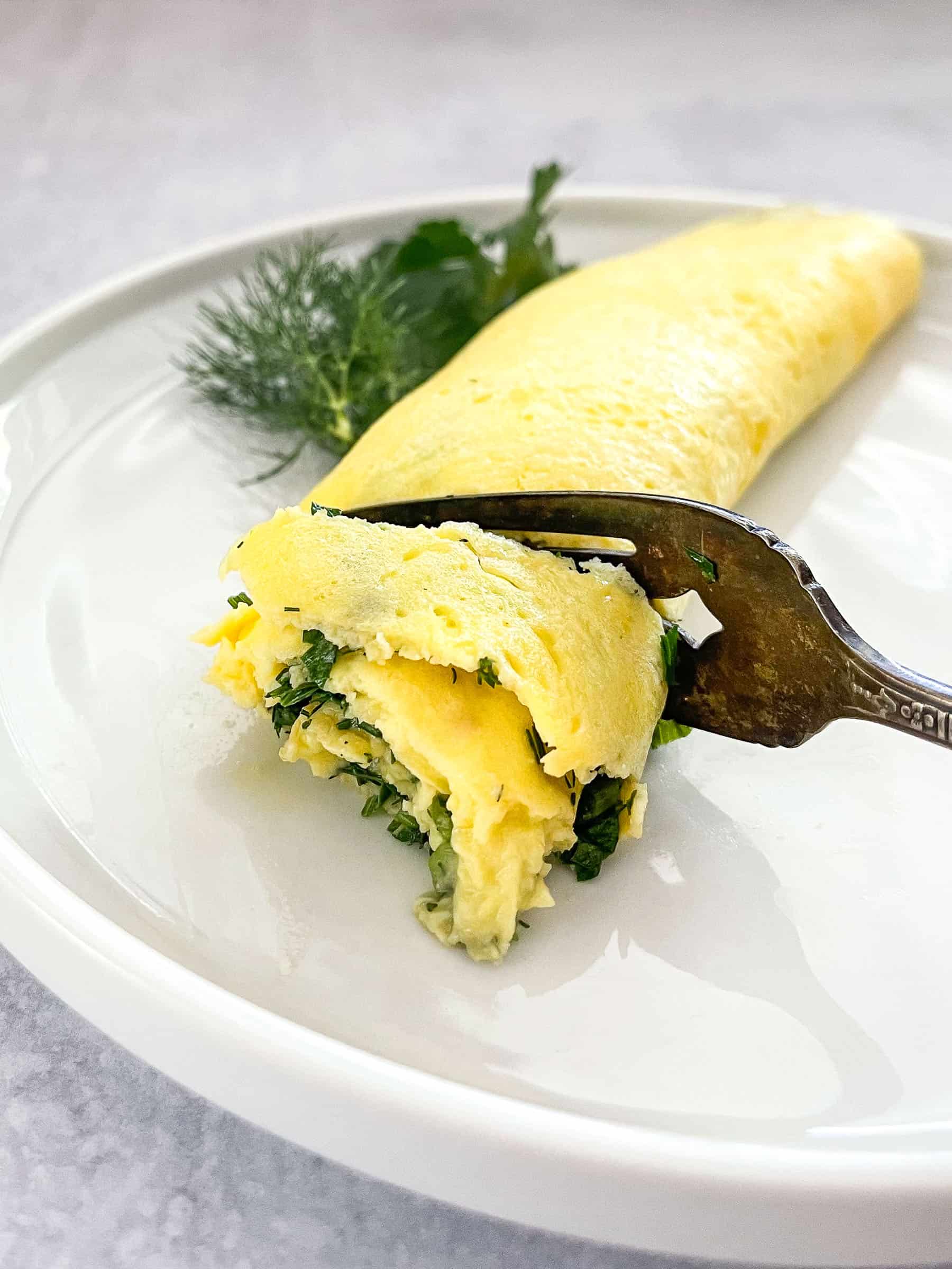 french omelet on white plate garnished with dill and parsley with antique fork cutting omelet