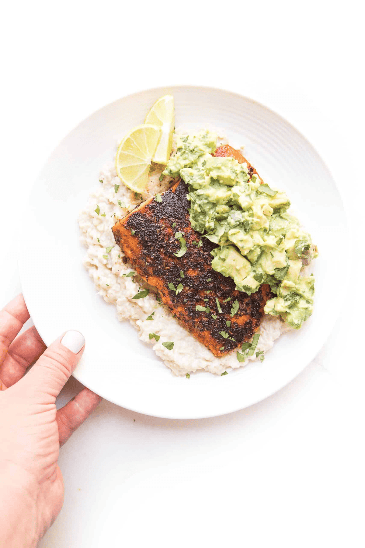 a hand holding a white plate with cauliflower rice, salmons, chunky guacamole and slices of lemon