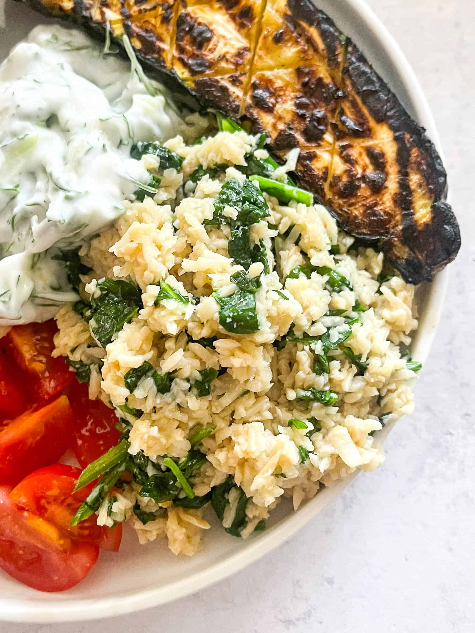 hearts of palm rice pilaf with spinach next to greek chicken kabobs, tomatoes, and tzatziki sauce on a white plate with a white background