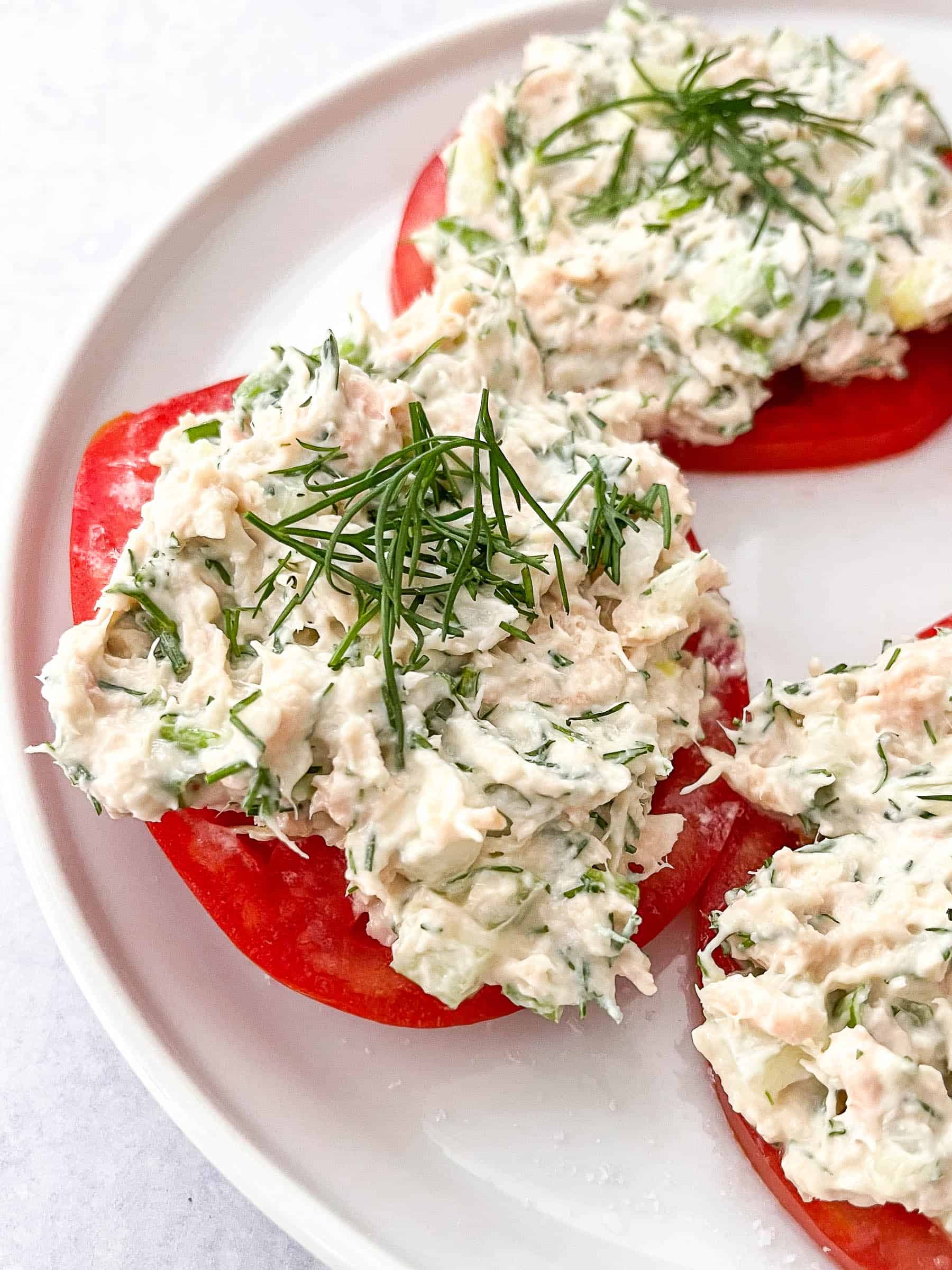 Tuna salad topped with dill on sliced tomatoes on a white plate