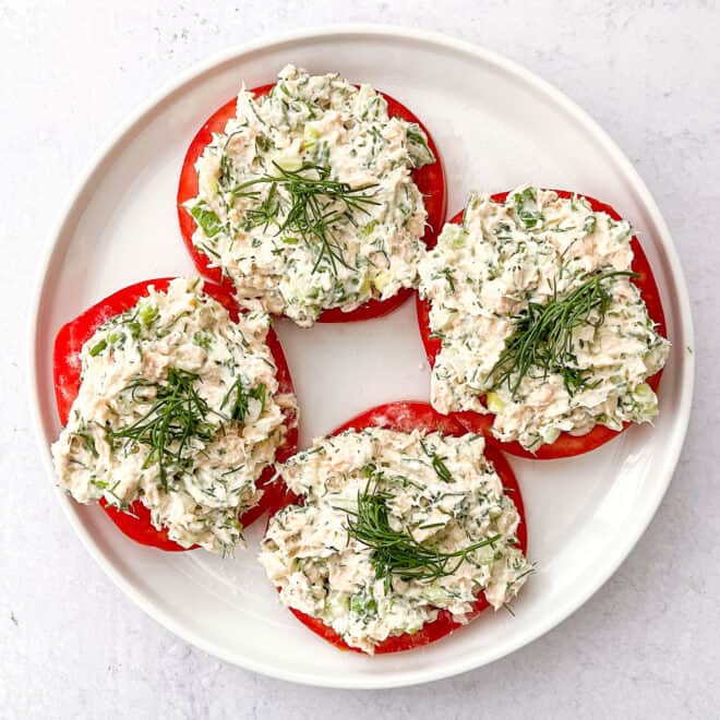 Tuna salad topped with dill on sliced tomatoes on a white plate