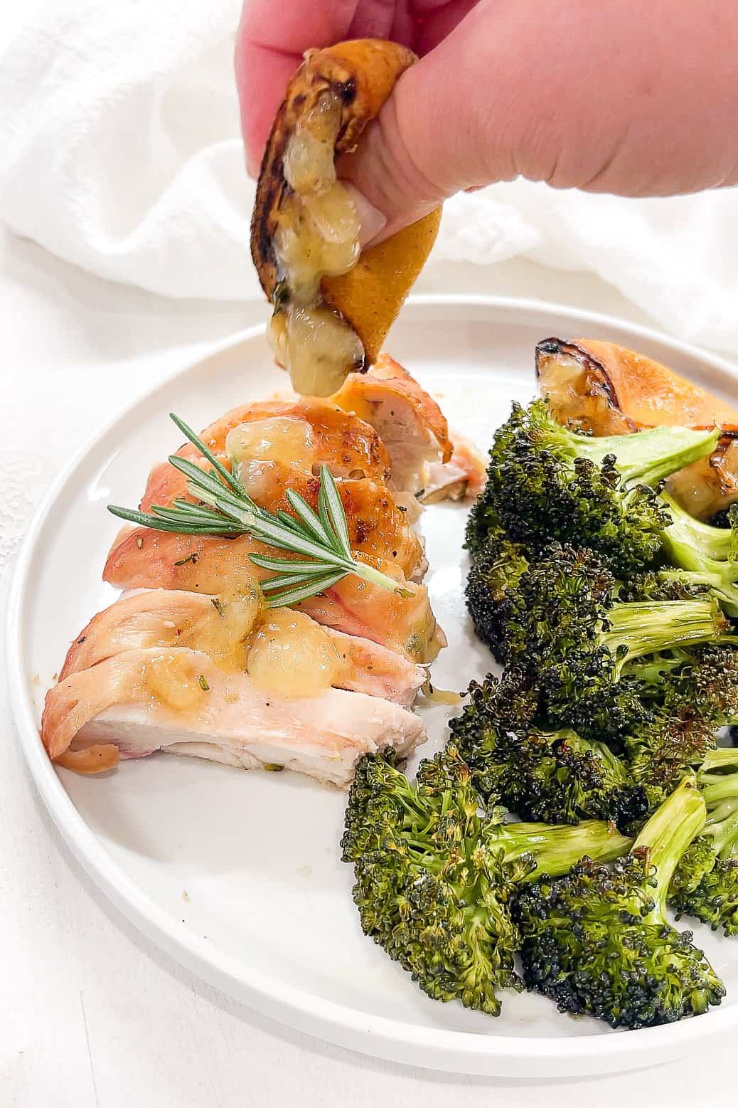 a hand squeezing a grilled lemon on top of sliced flattened chicken on a white plate next to a pile of roasted broccoli