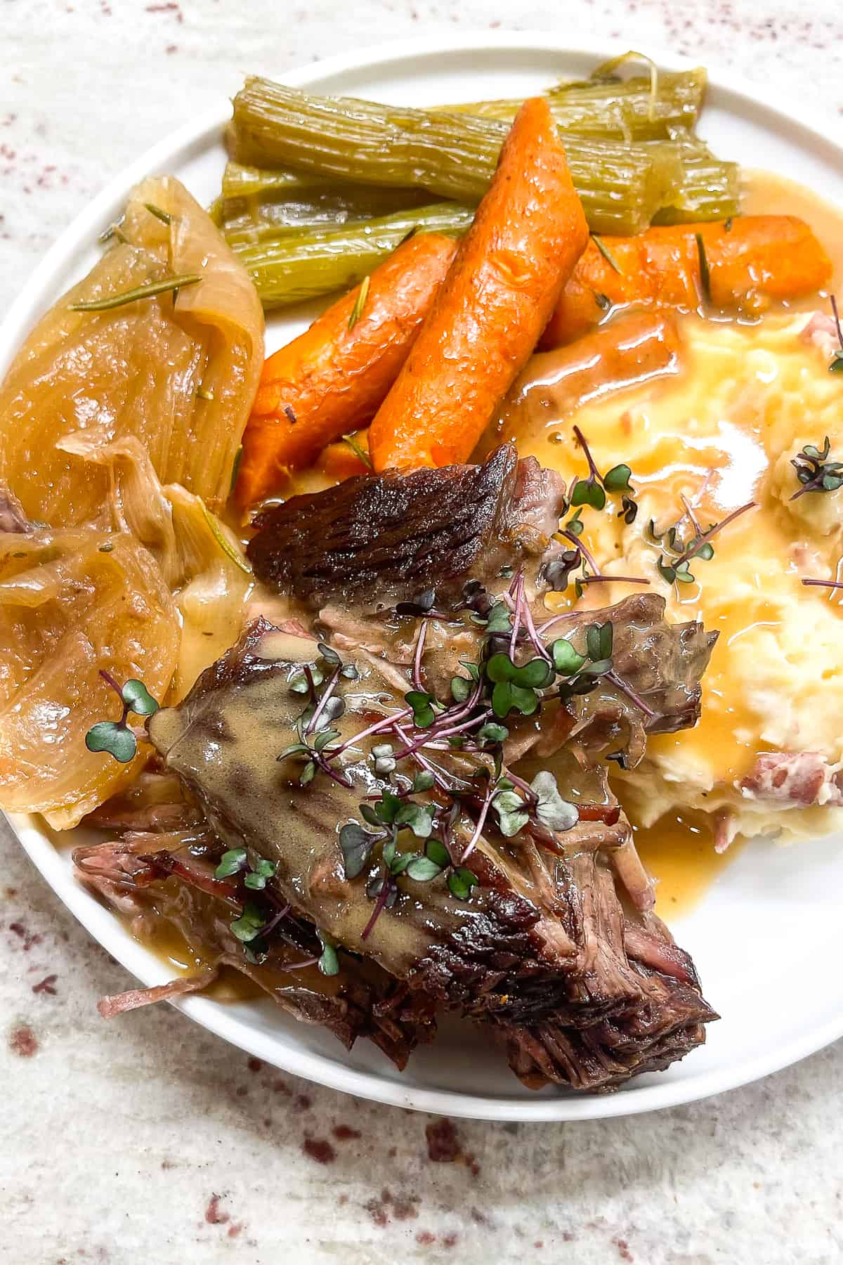 a close up of a cooked dutch oven pot roast topped with homemade beef gravy next to cooked onions, carrots, and celery