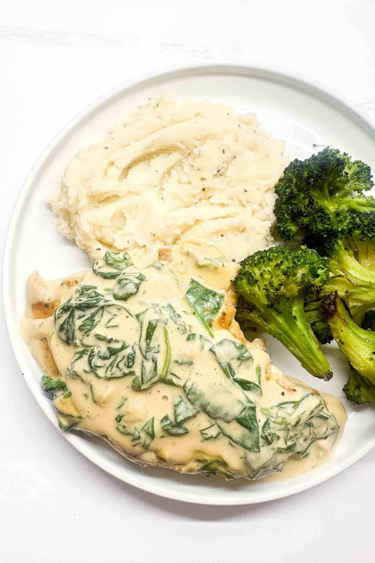 a photo showing a white plate filled with boursin chicken topped with spinach next to mashed cauliflower and roasted broccoli