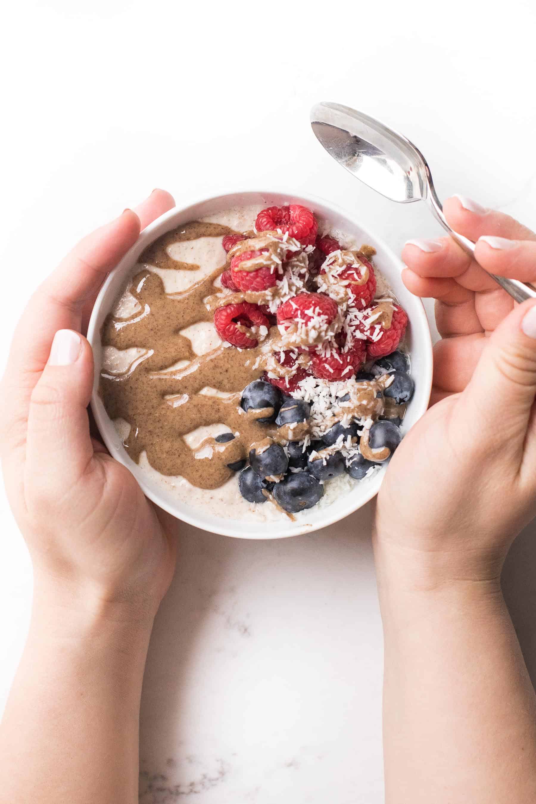 2 hands holding a bowl with breakfast porridge, berries and nut butter